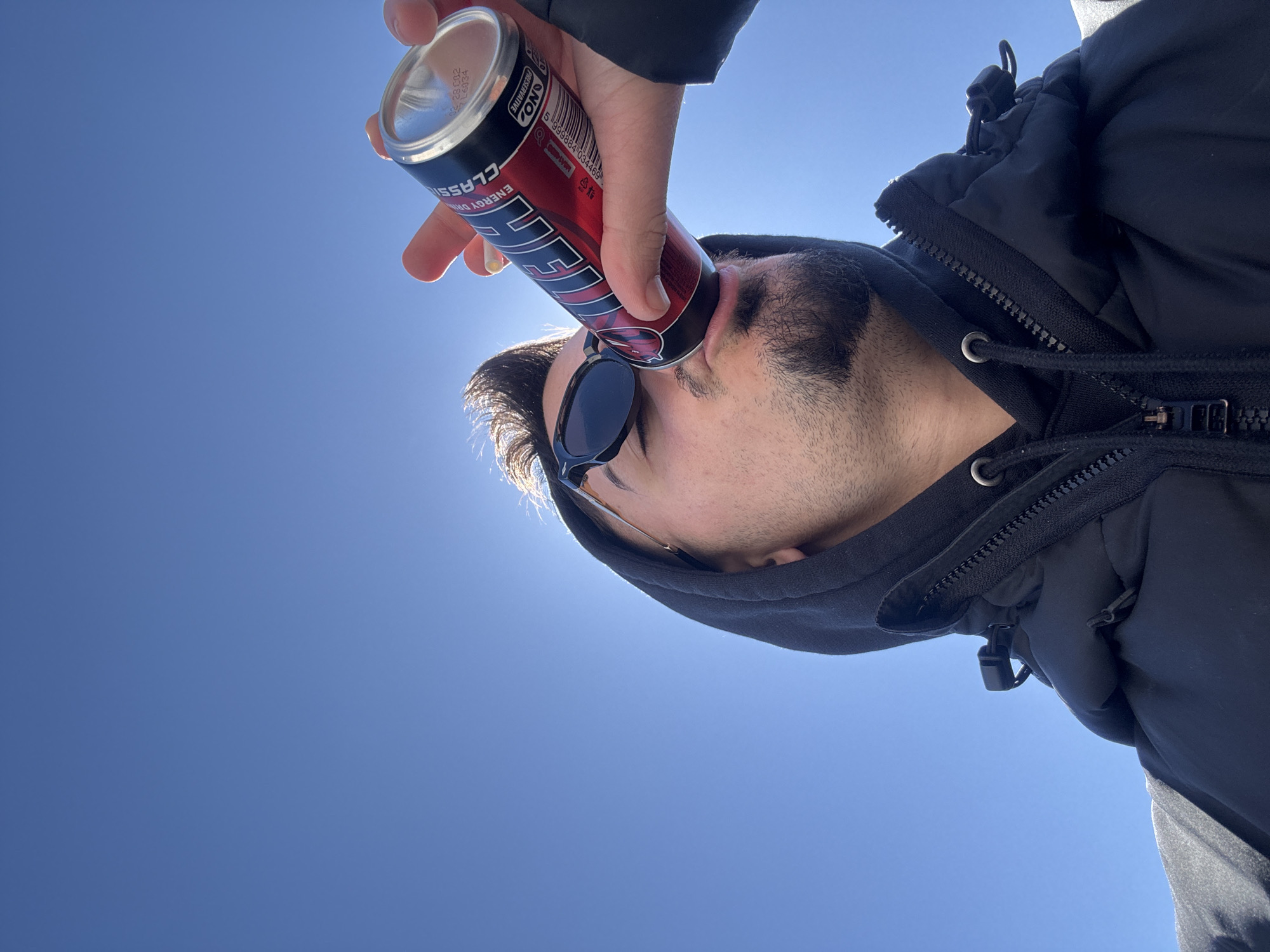 A man wearing a black hooded jacket and sunglasses is drinking from a red and black energy drink can against a clear blue sky. The brand name on the can is "HELL ENERGY DRINK CLASSIC". The location is not discernible, so I cannot state whether this image was taken in the city of Sofia, Bulgaria.