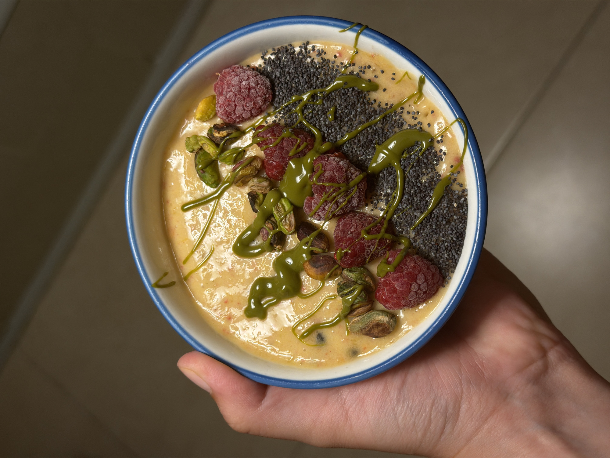 A hand is holding a blue-rimmed white bowl filled with a creamy, light orange smoothie. The smoothie is topped with several fresh raspberries, shelled pistachios, a scattering of black chia seeds, and a drizzle of vibrant green sauce, likely matcha or avocado. The background is blurred and appears to be indoors, with a dark, textured wall and a faint diagonal line suggesting a shelf or partition. The lighting is dim, casting shadows and highlighting the textures of the food. There is no discernible indication of a specific location in Tel-Aviv, Israel, in this image.