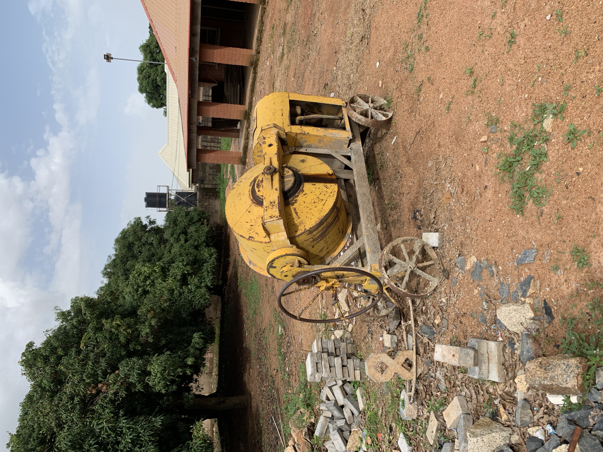 The image depicts an outdoor scene in the city of Jos, Nigeria. A yellow concrete mixer is positioned in the foreground, resting on a dusty, reddish-brown ground. To the left of the mixer, a pile of construction debris, including bricks and concrete pieces, is visible. In the background, a lush green tree dominates the left side of the frame, while a red-roofed building with brick pillars stands on the right. The sky above is partly cloudy. The environment appears to be a construction or renovation site.