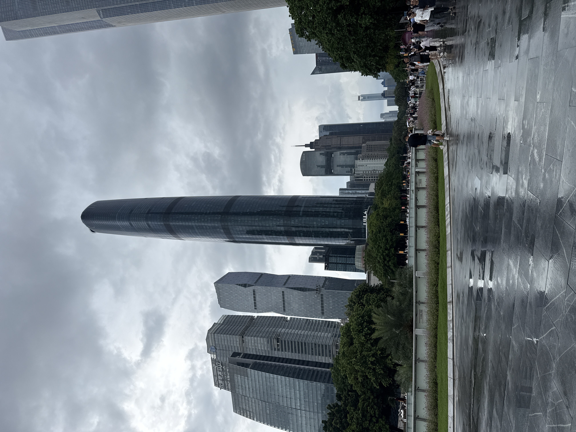 The image captures a contemporary urban scene in Tianhe District, China, likely during or shortly after a rain shower. The foreground features a dark, wet, stone-tiled plaza reflecting the overcast sky and the surrounding architecture. A scattering of people are visible walking along the plaza, with one individual in the mid-ground attending to a dog. Beyond the plaza, a manicured green lawn and flower beds are bordered by a decorative metal fence. In the background, a cluster of modern skyscrapers dominates the skyline, characterized by sleek glass facades and varied architectural designs. Notably, a tall, slender, dark-colored skyscraper stands prominently in the center, flanked by other significant buildings. Trees line the edge of the landscaped area, their foliage contributing to the urban greenery. The sky is heavily clouded, suggesting a diffused light or impending weather.