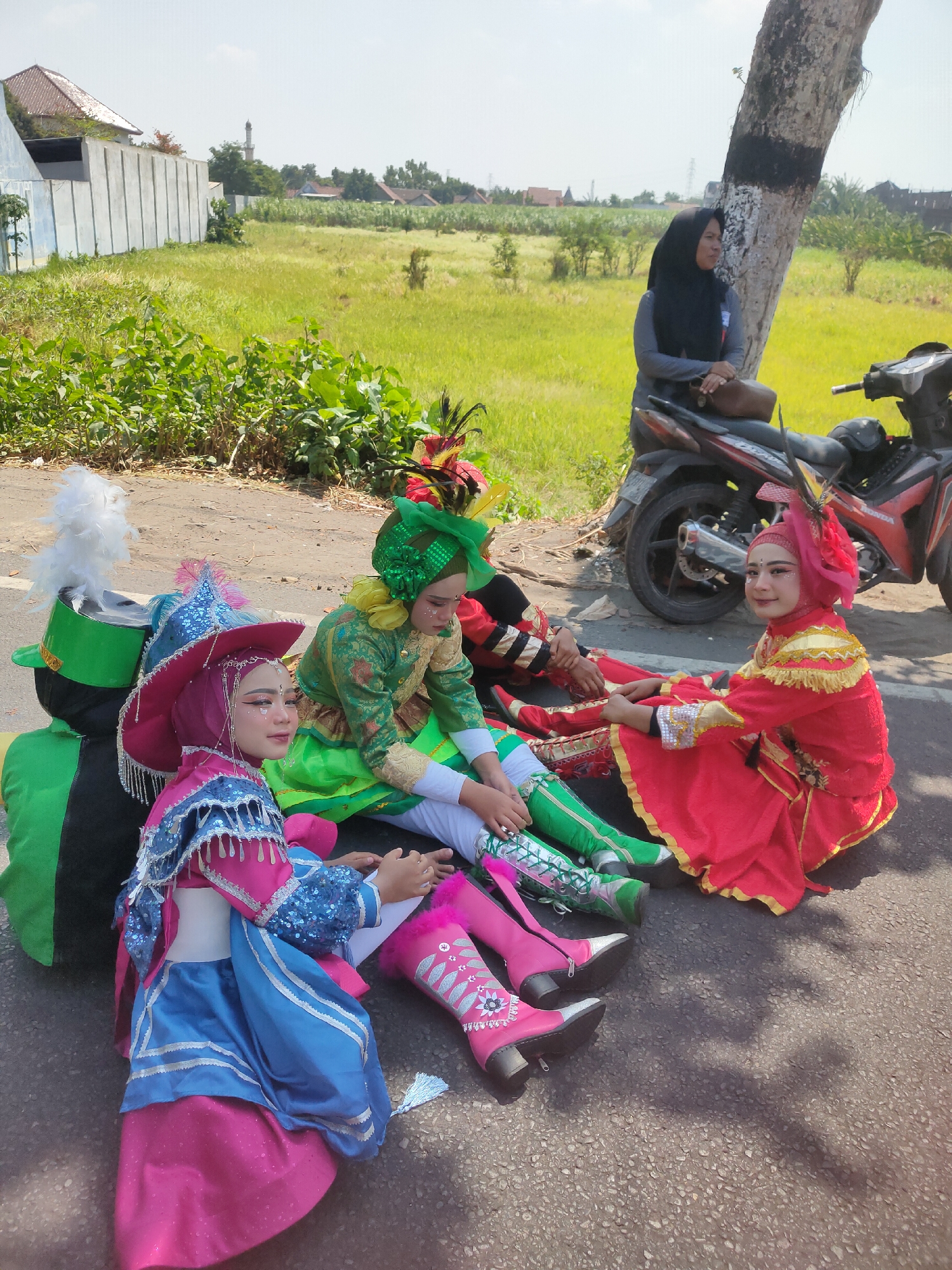 In Ngasem, Indonesia, a group of five young individuals, adorned in vibrant, traditional performance costumes, are seated on the asphalt road. The costumes feature a mix of colors including pink, blue, green, and red, with elaborate embellishments like sequins, tassels, and feathers. Two individuals wear tall, decorative boots. A motorcycle and a woman wearing a black hijab are visible in the background, to the right of the frame. The scene is set against a backdrop of lush green fields and a bright, clear sky, suggesting an outdoor event or parade.
