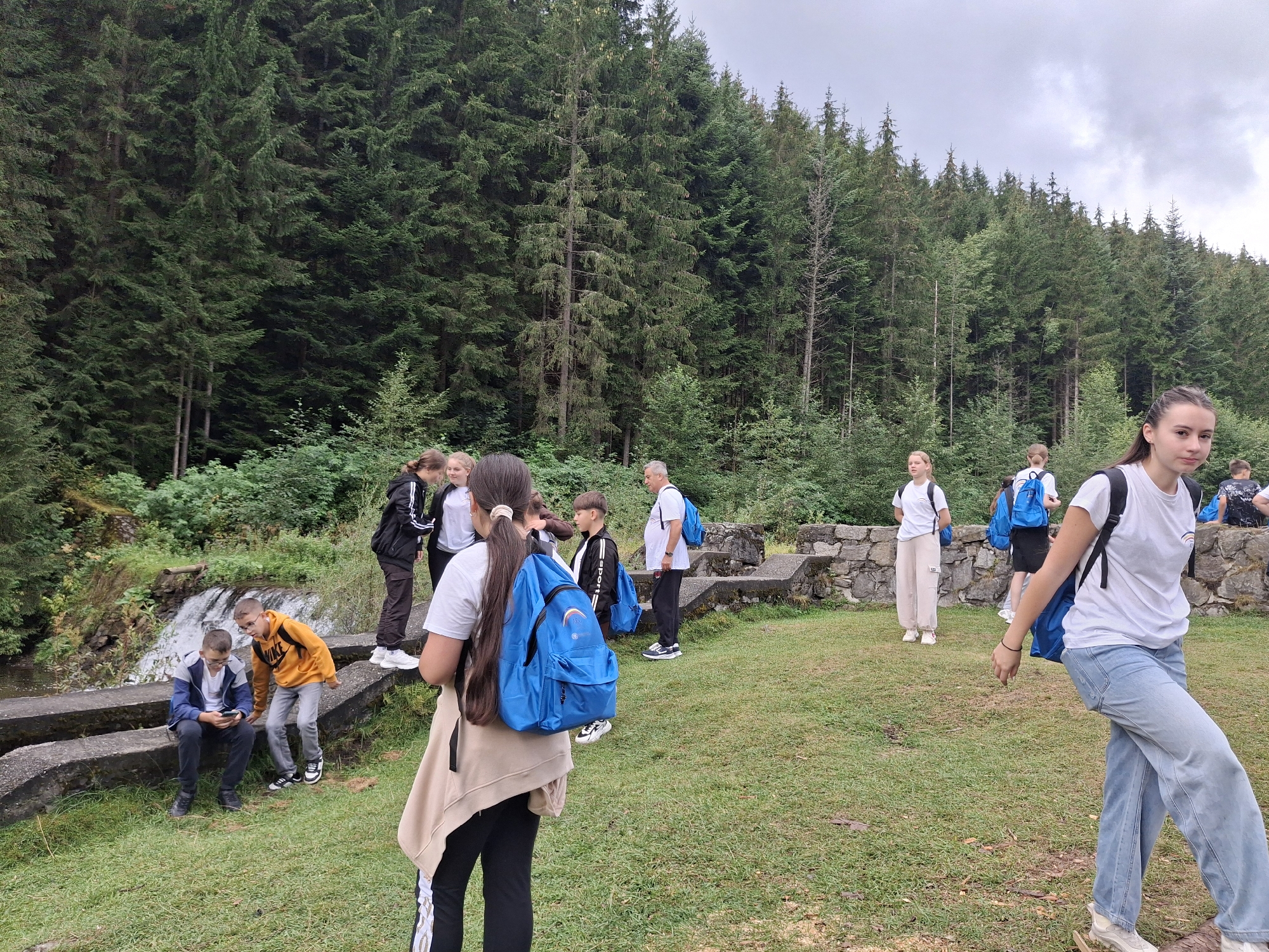The image depicts a group of people, primarily teenagers and a few adults, in an outdoor setting near a waterfall. The environment is characterized by a dense forest with tall evergreen trees, a grassy area, and some stone structures. A small waterfall cascades down rocks into a stream on the left side of the image. Several individuals are wearing backpacks, suggesting they are on a hike or excursion. One young man in an orange hoodie is looking at a smartphone, while another, in a blue jacket, sits beside him. A girl in the foreground, facing away from the camera, wears a white t-shirt and light-wash jeans, with a blue backpack. Another girl in a white t-shirt and light-colored pants is visible further back. An adult male in a white t-shirt and dark pants, also with a backpack, stands near some stone ruins. The sky is overcast. This scene appears to be located at the Valea Rece, a popular tourist spot in Sândominic, Romania, known for its natural beauty and waterfalls.