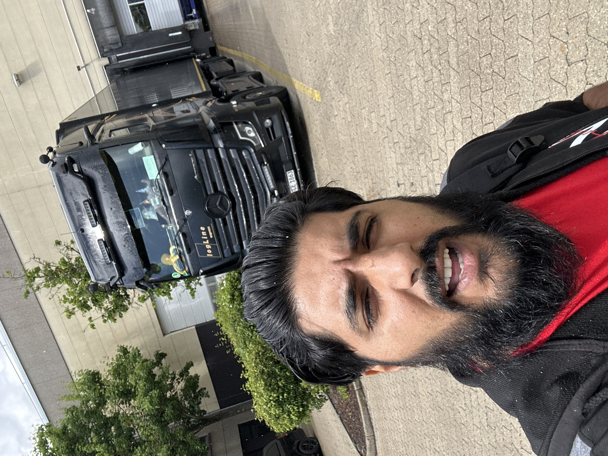 A man with dark hair and a beard is taking a selfie in front of a black semi-trailer truck. The truck has "logLine express" written on the side and a Mercedes-Benz logo on the front. The license plate is visible and reads "CE 1863". The man is wearing a black jacket over a red shirt. He is standing on a paved area with yellow lines. In the background, there is a large building with a loading dock and trees. The image appears to have been taken outdoors, possibly at a logistics facility. There are small white particles visible in the air, suggesting it may be raining or snowing lightly. The location is identified as Schweitenkirchen, Germany.