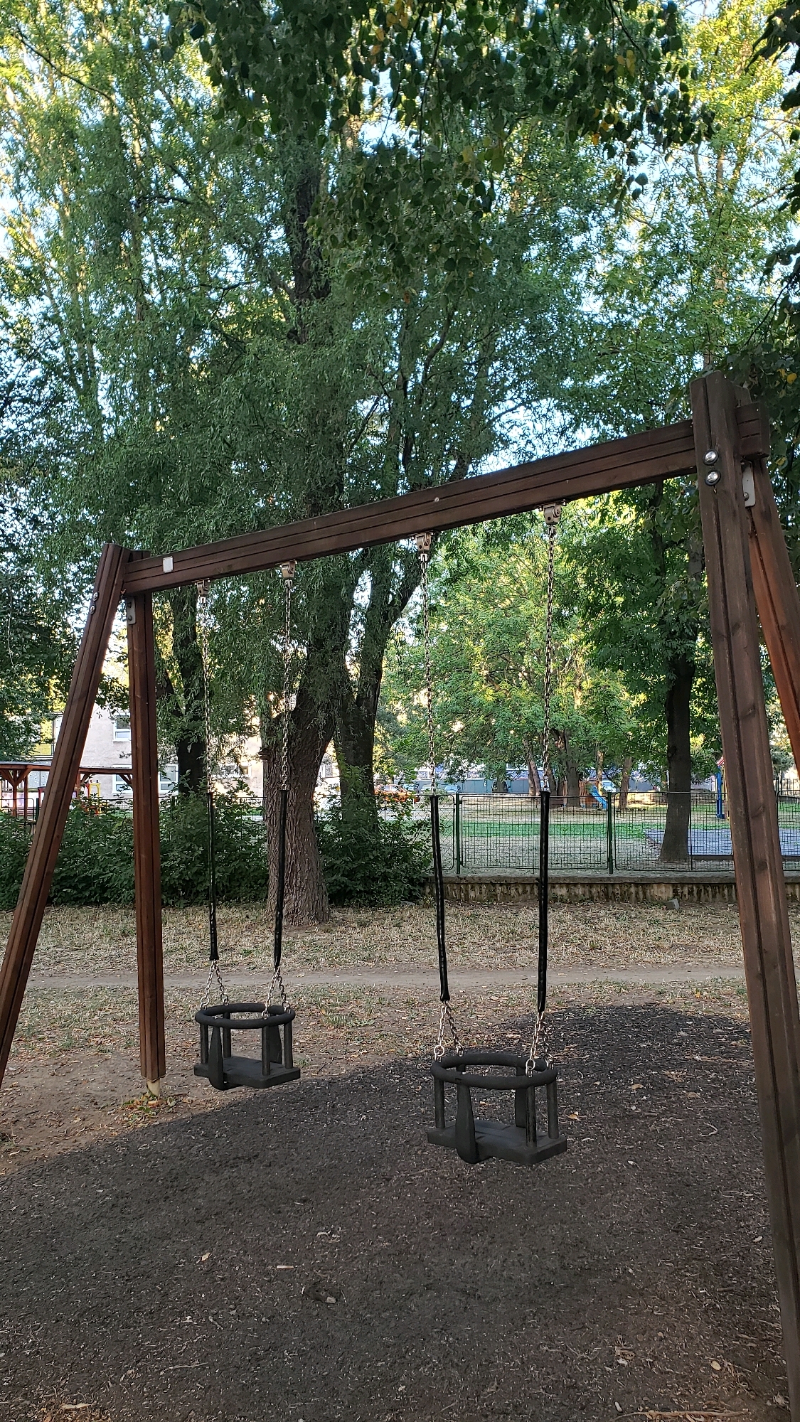 The image shows an empty swing set with two black baby swings, suspended from a wooden frame. The swings are hanging over a dark, soft surface, likely intended as a safety measure for a playground. In the background, lush green trees create a canopy, with some buildings visible through the foliage. A fence separates the playground from a path and grassy area beyond. The scene appears to be in a park or residential area, with the afternoon sun casting long shadows. The specific location in Prešov, Slovakia, is an outdoor playground.