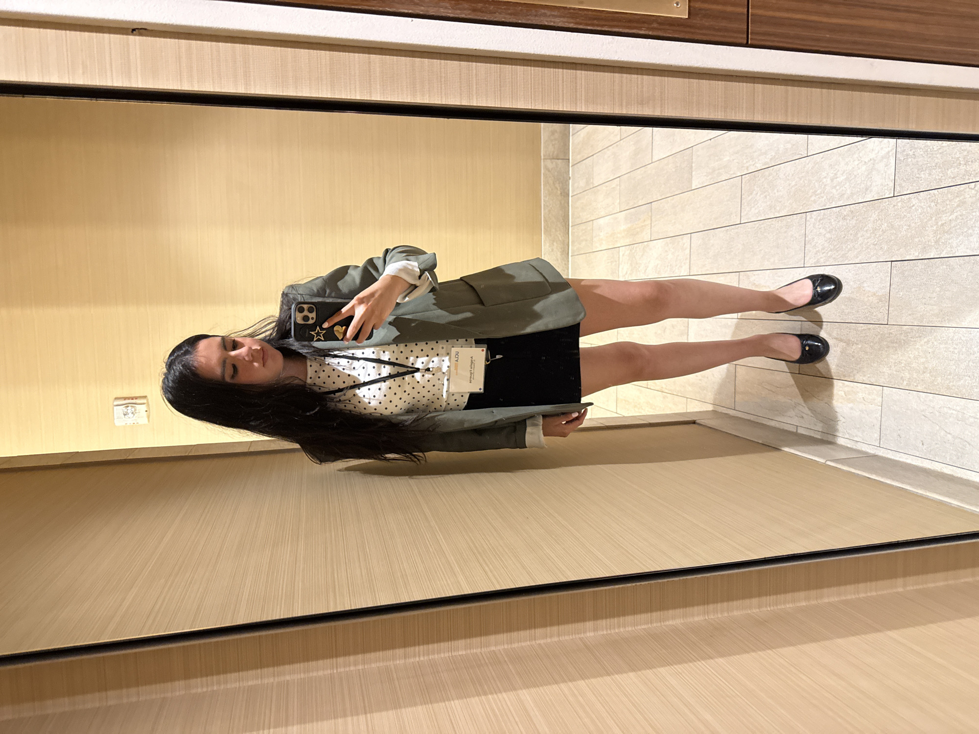 A young woman with long, dark hair is taking a mirror selfie in a hallway with wood-paneled walls and beige floor tiles. She is wearing a white polka dot shirt, a dark skirt, a green blazer, and black ballet flats. A lanyard with a name tag is visible around her neck. The overall scene suggests a hotel hallway in a conference center or similar venue. The image is taken from the chest down. There is no indication of the specific location in Los Angeles, United States of America.