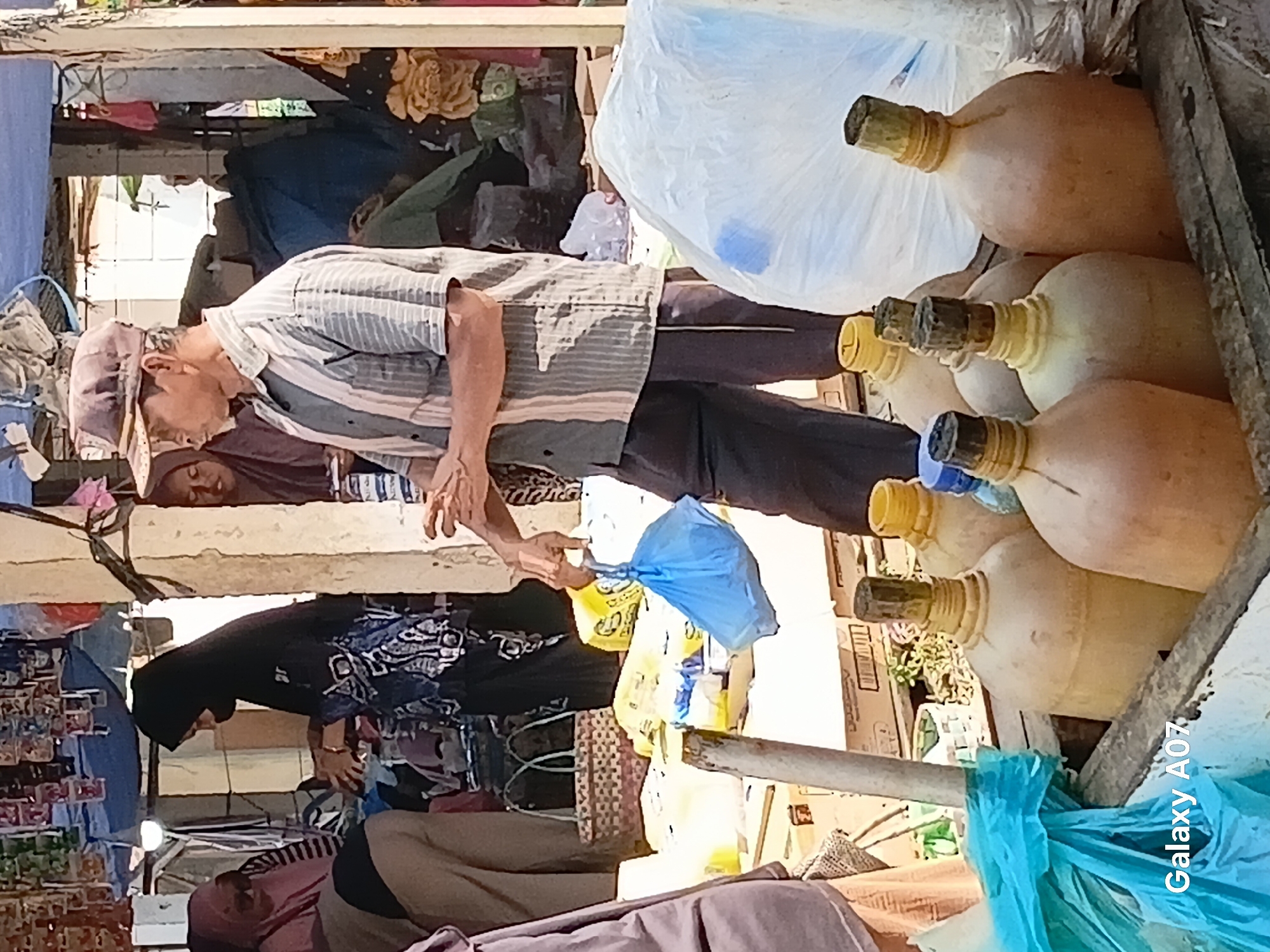 A medium shot captures an outdoor market scene in Margo Rukun, Indonesia. In the foreground, a man wearing a striped shirt, dark pants, and a brimmed cap is seen from the side as he holds a blue plastic bag. In the mid-ground, several large, tan-colored plastic jugs with yellow caps are visible, some appearing to be partially filled. Behind the jugs, more market stalls and people are visible. To the left, a woman in a pink headscarf is interacting with another person partially obscured. Further back, a person in a black hijab is visible. The environment appears to be a bustling market with goods displayed on tables and shelves, covered by tarps in some areas. The lighting suggests it is daytime.
