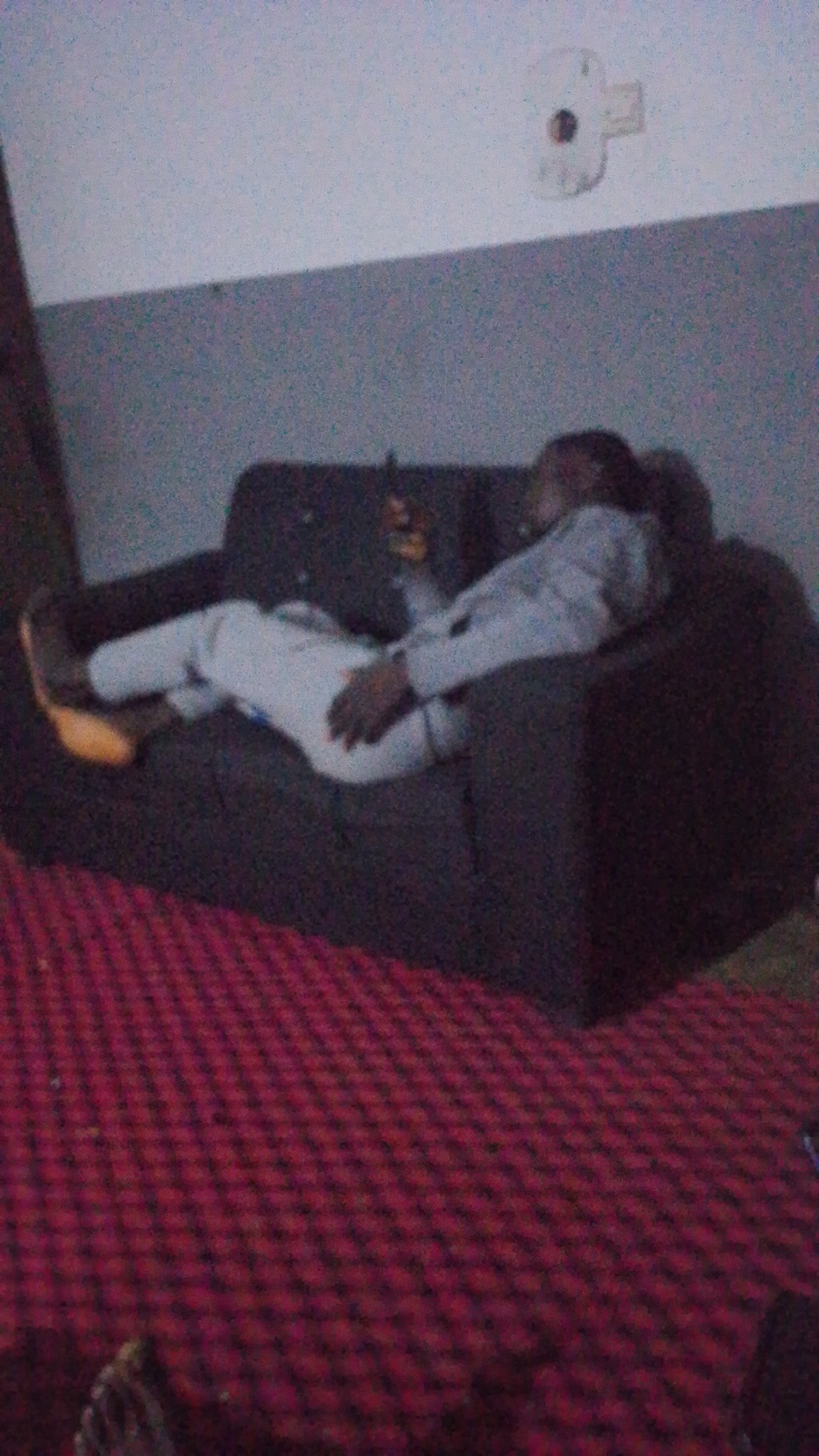 A person is lying on a dark couch in a dimly lit room, with their legs extended and their head propped up. They are wearing white pants and a light-colored shirt or jacket. Their left hand is resting on their lap, and their right arm is bent with their hand near their face. A dark-colored phone is visible in their right hand. The couch is situated on a red and black patterned carpet. The wall behind the couch is a dark gray, and the ceiling is white. A white object with a dark circular center is mounted on the white ceiling. The overall lighting is low, suggesting it might be nighttime or the room is not well-lit.