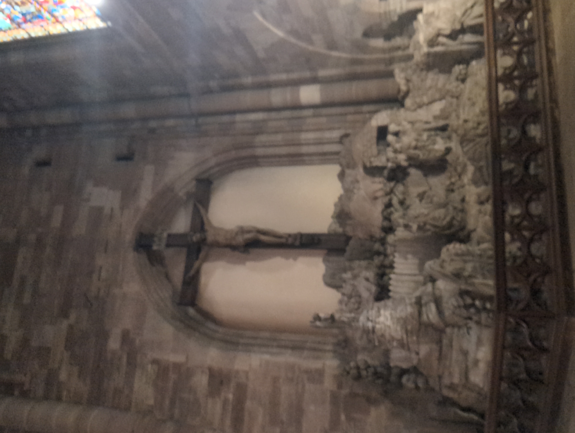 This image depicts a large crucifix with a statue of Jesus Christ positioned in an alcove within a stone church interior. The crucifix is dark wood and appears to be very old. Below the crucifix, there is a detailed stone sculpture depicting a scene with multiple figures, possibly representing a biblical event. The figures are carved in relief and appear to be in various stages of distress or contemplation. The architecture of the church is visible, with large stone blocks forming the walls and arches. A stained-glass window can be seen in the upper right corner, admitting light into the space. In the foreground, there is a wooden railing with intricate carvings. The overall impression is one of solemnity and historical significance. This scene is located in the Reims Cathedral, also known as the Cathedral of Notre-Dame of Reims, in Reims, France.
