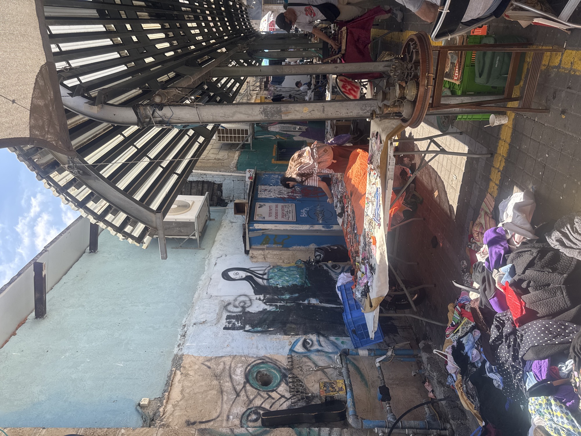The image depicts a street scene in Tel Aviv, Israel. The location appears to be a market or a flea market, with vendors selling goods on tables and the ground. The scene is characterized by narrow passageways between the vendors.
Several individuals are visible, including vendors and shoppers. The goods on sale appear to include clothing, jewelry, and other assorted items. There is evidence of graffiti on the walls of the buildings along the street. The street is covered by an open metal awning.
