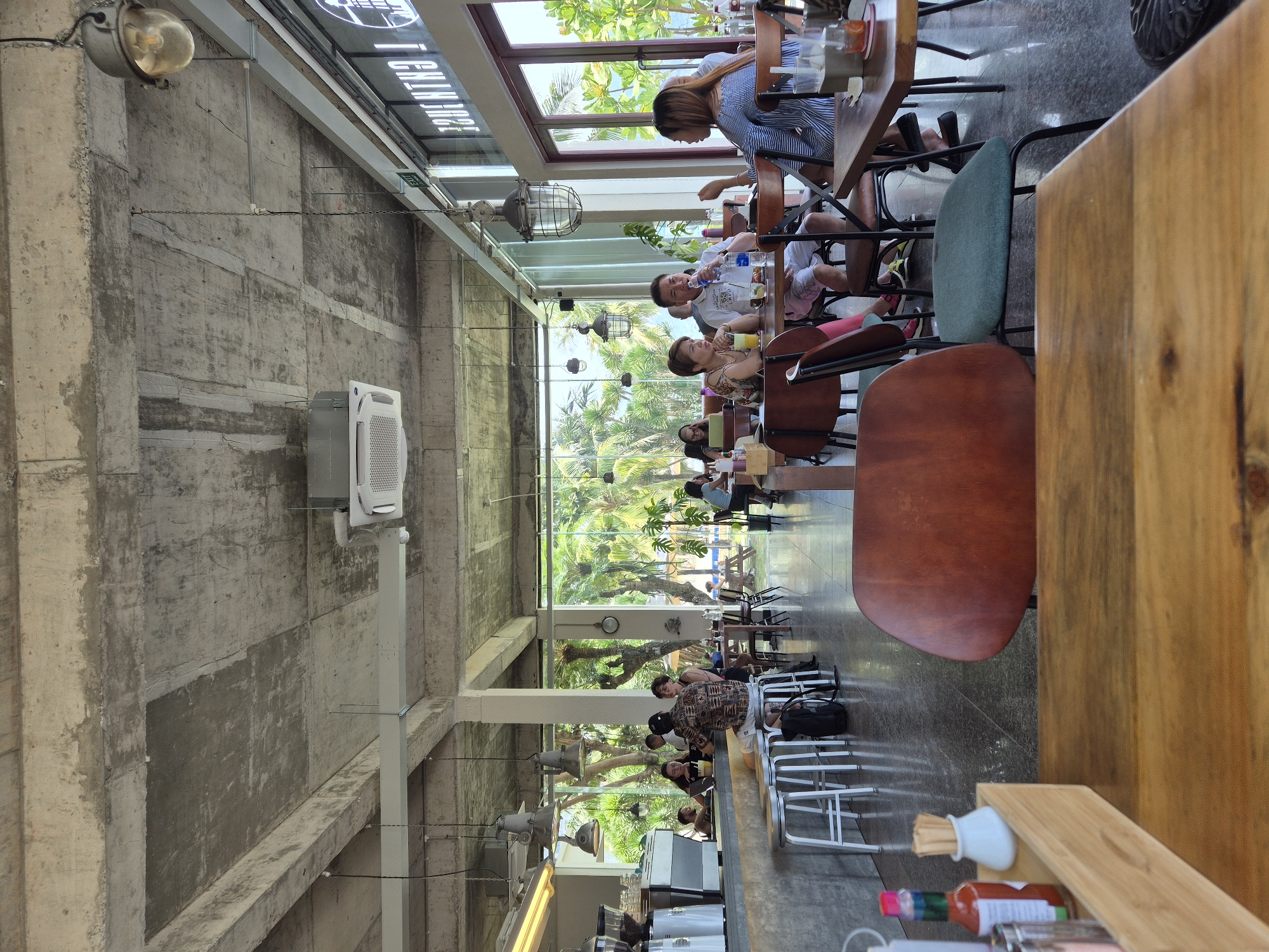 The image depicts the interior of a contemporary restaurant or cafe with an industrial-chic aesthetic, located in Đà Nẵng, Vietnam. The space features an exposed concrete ceiling with visible beams and ductwork, complemented by hanging industrial-style light fixtures. Large windows offer a view of lush tropical greenery and a body of water, suggesting a beachfront or poolside location.
In the foreground, a wooden table occupies the lower portion of the frame, with a close-up view of a wooden tray containing condiments like ketchup and toothpicks in a small white holder. Two chairs are partially visible, one with a rich brown leather seat and back, and another with a teal fabric seat and black metal frame.
The mid-ground shows patrons seated at tables and at the bar. A prominent bar area features a concrete counter with several white stools, where a few individuals are seated. Further into the restaurant, groups of people are engaged in conversations and enjoying meals or drinks. One table in the center has a group of four people, two men and two women, seated. A man in a white t-shirt and patterned shorts is drinking from a bottle, and a woman in a striped top is gesturing with her hand. Another couple is seated to their left, with a woman in a floral top and a man looking in their direction.
In the background, beyond the interior seating area, more tables and chairs are visible, with patrons further scattered. The large windows reveal palm trees and other dense vegetation, and glimpses of the ocean or a large pool. The overall atmosphere appears relaxed and airy, with ample natural light flooding the space. An "EXIT" sign is visible above a doorway. The signage on the far right wall, partially visible and reversed, reads "I CAN'T JUST".