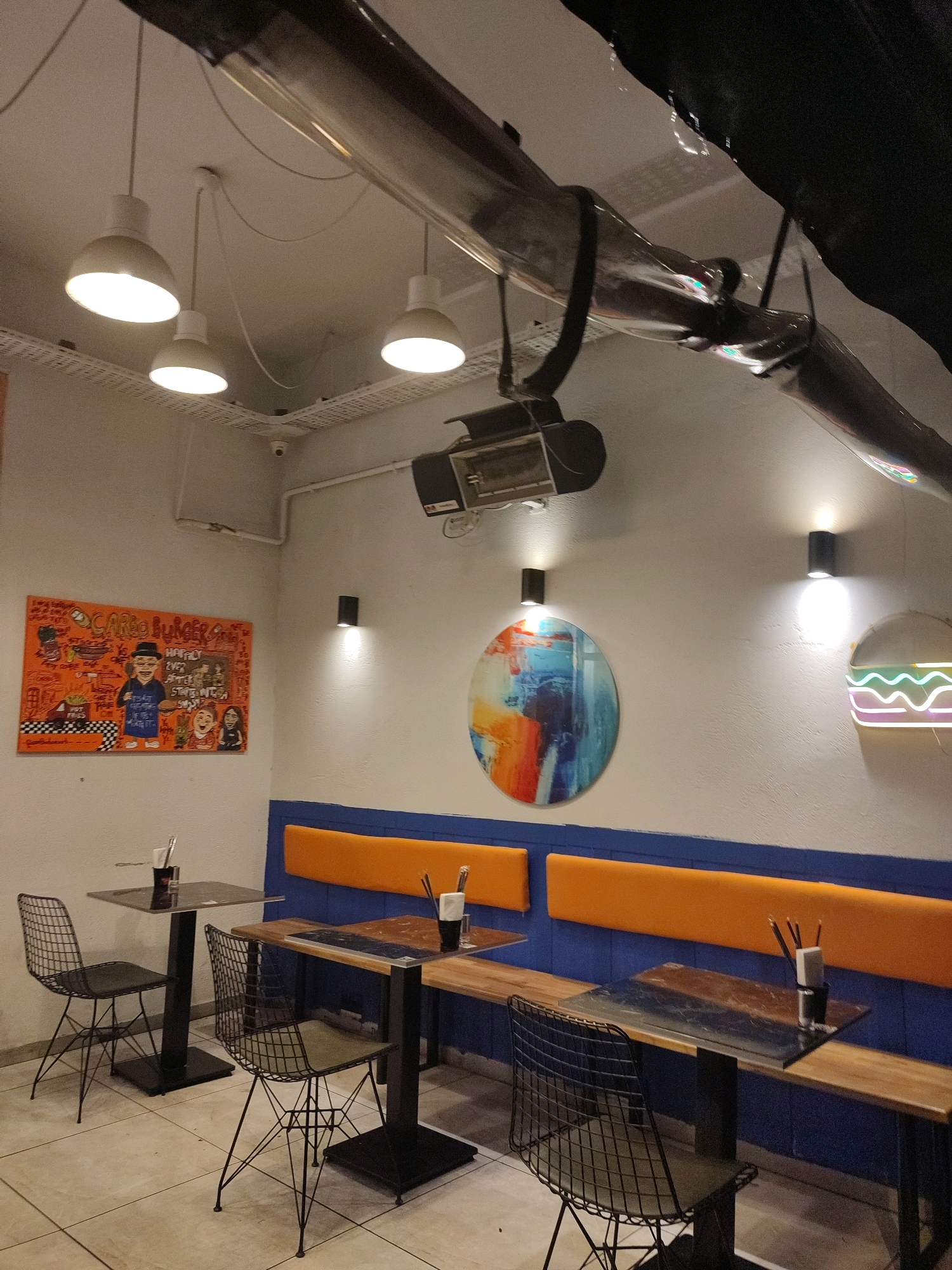The image shows the interior of a restaurant with a modern and industrial design. The walls are painted white, with blue and orange cushioned seating along one wall. There are several tables with black metal chairs. On the wall, there is a colorful painting of a burger and people, and a circular abstract painting. There is also a neon burger sign. The ceiling has exposed pipes and lighting fixtures. The floor is tiled. No people are visible in the image. The image is taken from a slightly low angle, looking across the seating area. The restaurant appears to be located in Kadıköy, Turkey, as indicated by the "CARGO BURGER" sign, which is a known restaurant chain in Istanbul.