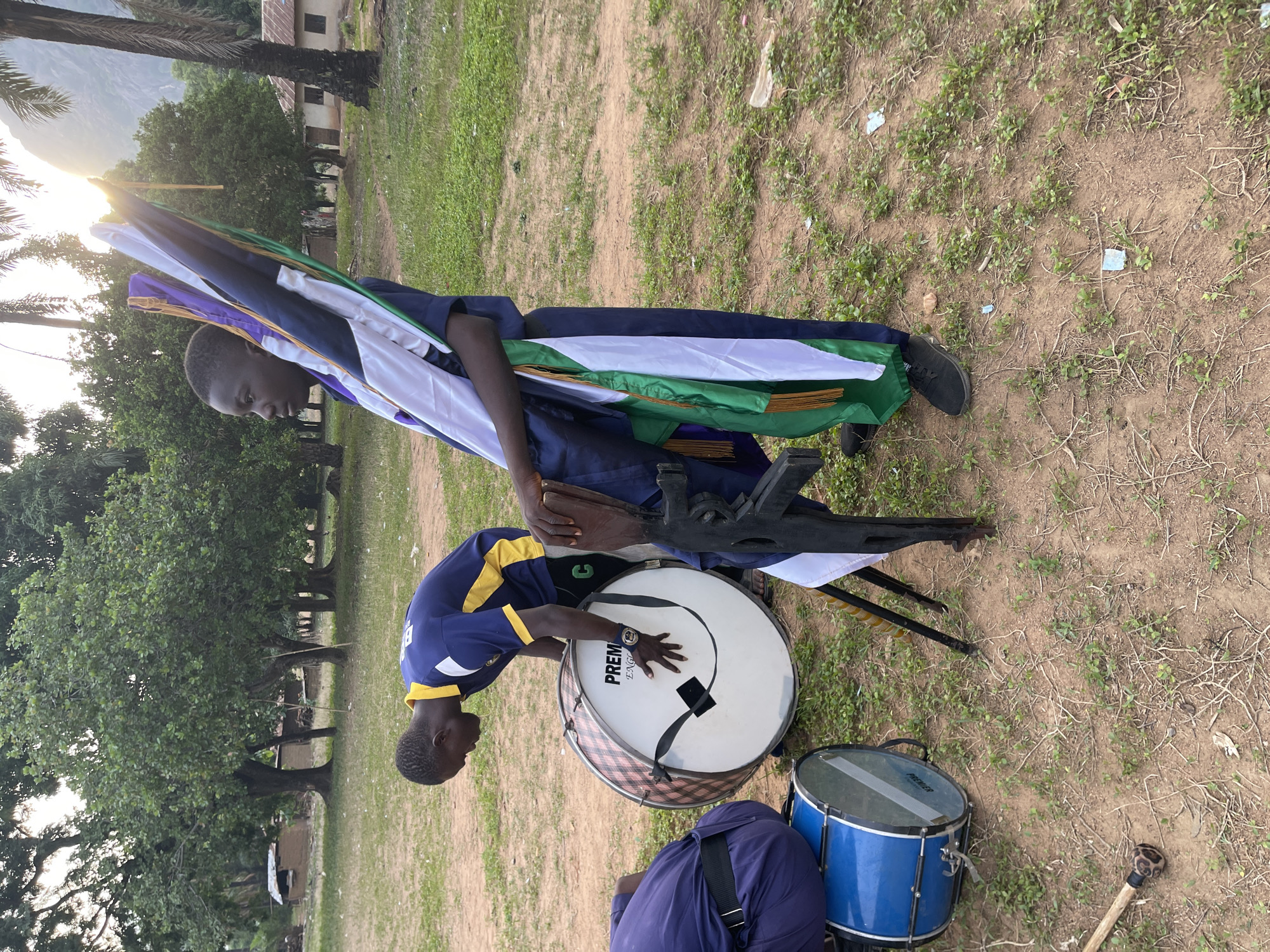 The image depicts an outdoor scene in what appears to be a grassy area, possibly a field or park, in Andaha, Nigeria. Two young men are present. One man is standing, holding a collection of banners or flags with different colors, including purple, white, and green. The other is bent over a large, white bass drum, apparently adjusting or tuning it. Beside the bass drum, there is a blue snare drum on the ground, and a drumstick is near the base of the blue drum. The standing man is wearing a dark blue outfit, while the other man is wearing a blue and yellow top. The ground is dry and covered with sparse grass. In the background, there are trees and what appears to be a building. The image has been rotated 90 degrees, with the horizon visible at the top, including a glimpse of a mountain and the sun.