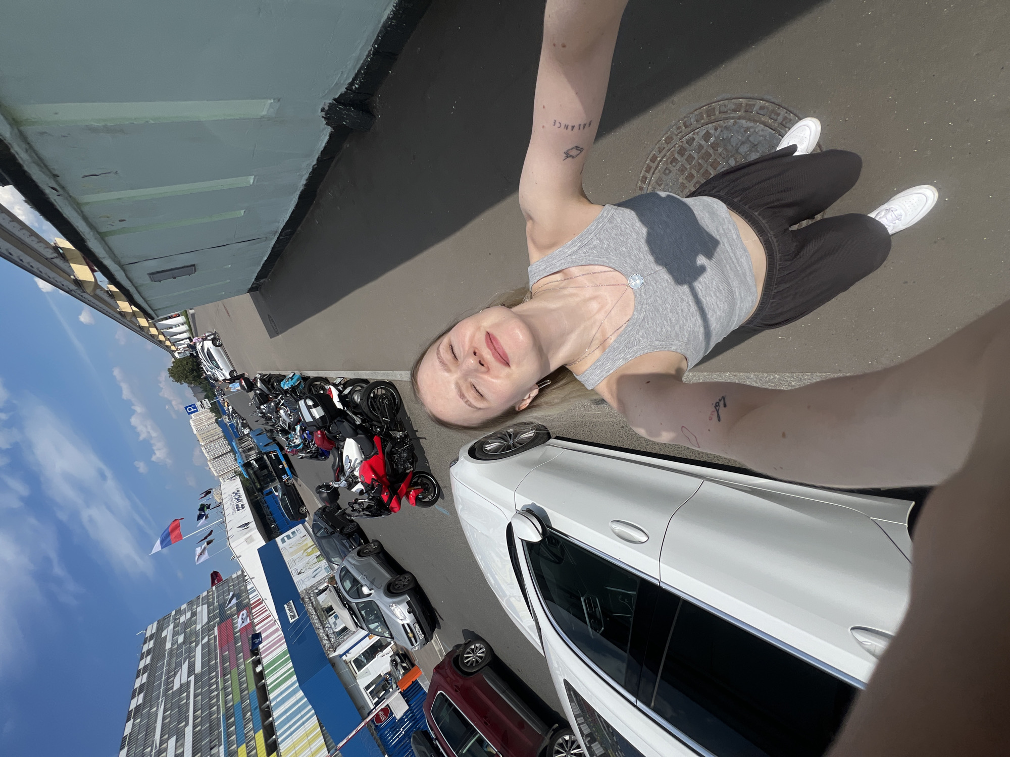 A woman with blonde hair is taking a selfie in a parking lot on a sunny day. She is wearing a grey tank top and black pants. Behind her, there are several cars and motorcycles parked. In the background, there is a modern building with a colorful facade, and Russian flags are flying from a pole. The scene is set in the city of Khimki, Russia.