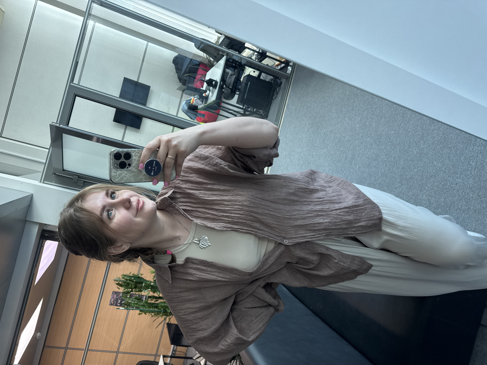A young woman captures a mirror selfie indoors, holding an iPhone equipped with a PopSocket that displays the text "Peace & Love EveryDay". She has light brown hair, blue eyes, and wears light pink nail polish and a gold ring. Her outfit includes a loose, textured, light brown button-up shirt worn open over a light cream tank top, paired with light-colored, possibly off-white, wide-leg pants or a skirt. A distinctive silver, heart-shaped pendant hangs from her necklace.
The setting appears to be a modern indoor space, likely an office building, lobby, or public area, characterized by large glass panels that reflect the surrounding environment. These reflections reveal wooden wall paneling, gray flooring or carpet, several office chairs and tables, computer equipment, and a green potted plant. The lighting suggests it is daytime. The woman maintains a neutral to slightly pleasant expression while looking at her phone.