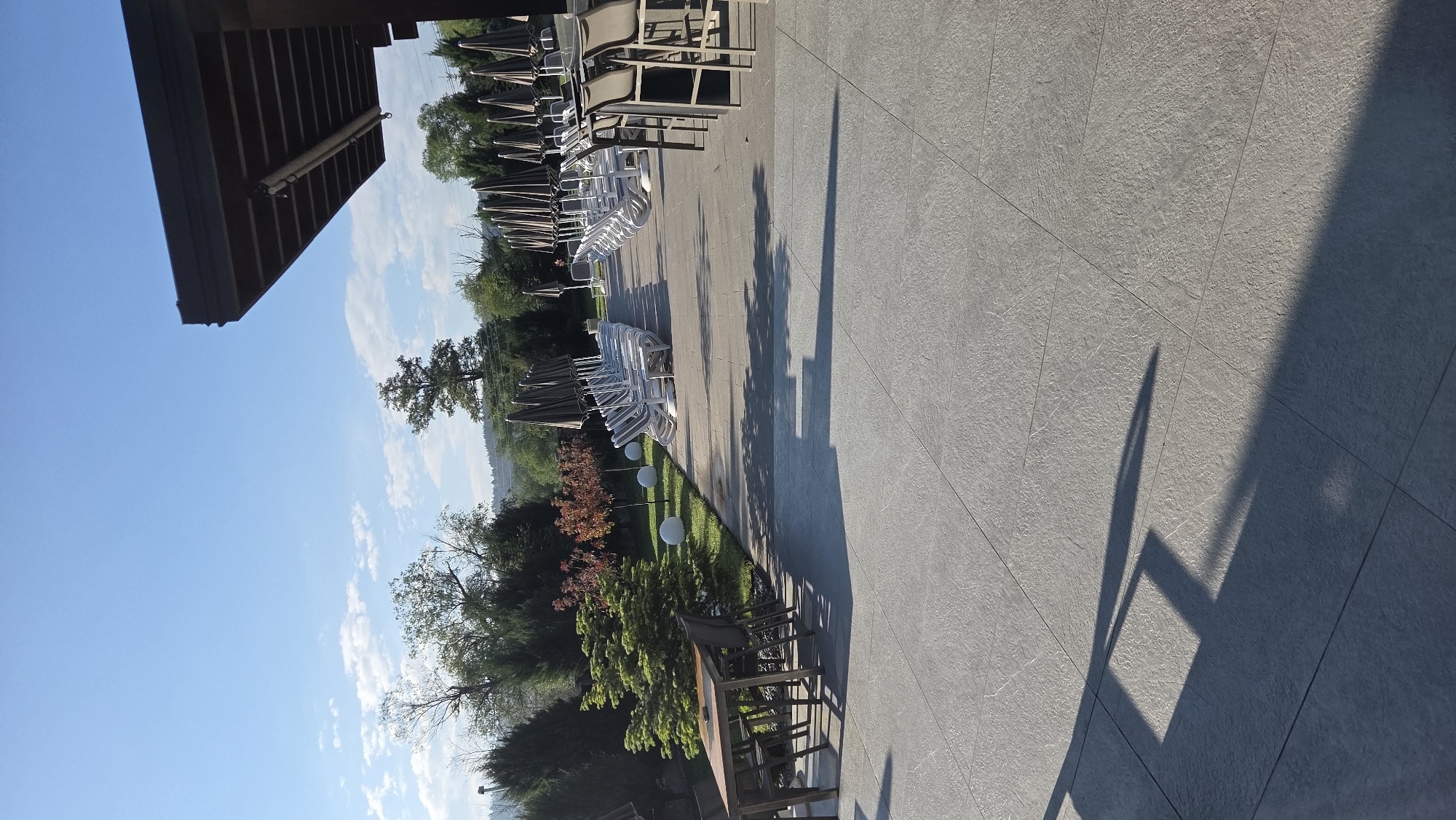 The image depicts an outdoor patio area in an unknown city. The patio is constructed with large, gray tiles and features rows of lounge chairs and umbrellas. The chairs are arranged in two groups: one with darker frames and another with white frames. Shadows cast across the patio suggest a sunny day. Adjacent to the patio, there is a landscaped area with trees and bushes. A wooden table and chairs are situated near the edge of the patio, close to the greenery. In the background, there are more trees, rolling hills, and a blue sky with scattered clouds. A building overhang is visible in the top-left corner of the image.