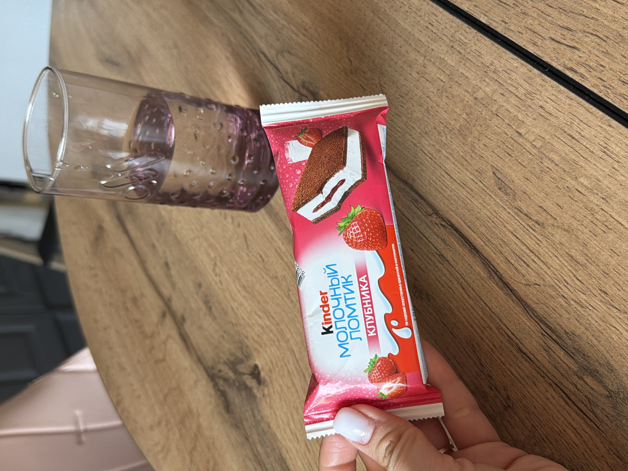 The image shows a partially filled glass and a Kinder Milk Slice bar, strawberry flavor, on a wooden surface. The bar is held by a person's hand with white nail polish. The text on the packaging is in Russian. The environment appears to be indoors on a wooden table or counter. The location is not discernible from the image.