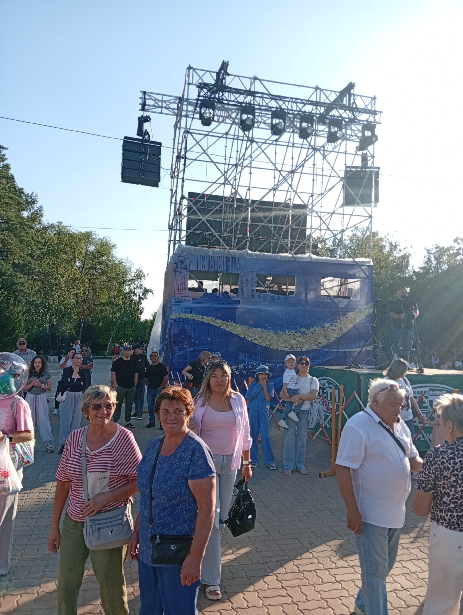 The image depicts an outdoor event, likely a concert or festival, taking place in the city of Kostanay, Kazakhstan. A large metal truss structure, equipped with speakers and lighting, dominates the background. In front of the stage, a blue banner with a design resembling a cityscape is visible. A crowd of people, varying in age and attire, are gathered. Some are walking, others appear to be observing the setup, and a few are actively engaged with each other. The ground is paved with interlocking stones. The overall atmosphere suggests a sunny day with clear skies. The presence of a camera operator with a tripod indicates that the event is being filmed.