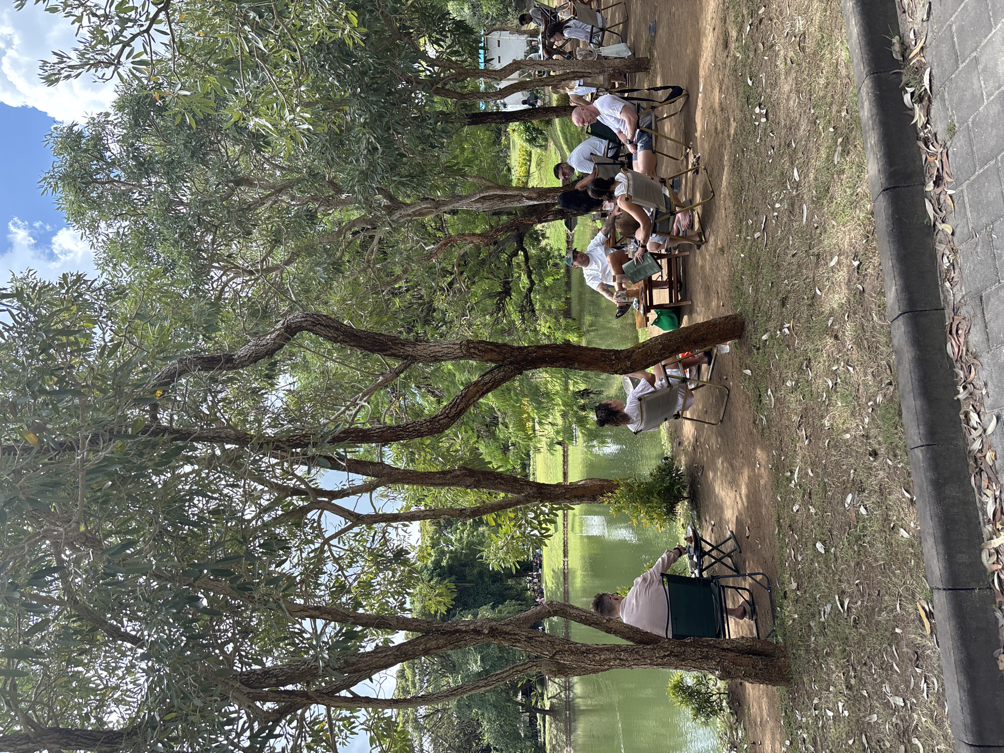 Captured at a park in Kuta Selatan, Indonesia, the image shows a gathering of people seated in chairs beside a body of water, likely a pond or small lake. Trees frame the scene, with branches extending across the top portion of the image and trunks visible in the foreground. Individuals are engaged in various activities, some interacting with others, while others appear to be observing the water or engaged in personal activities. The ground is covered with dirt, leaves, and grass. A paved walkway is on the far right. The overall setting appears relaxed and informal, indicative of a park environment.