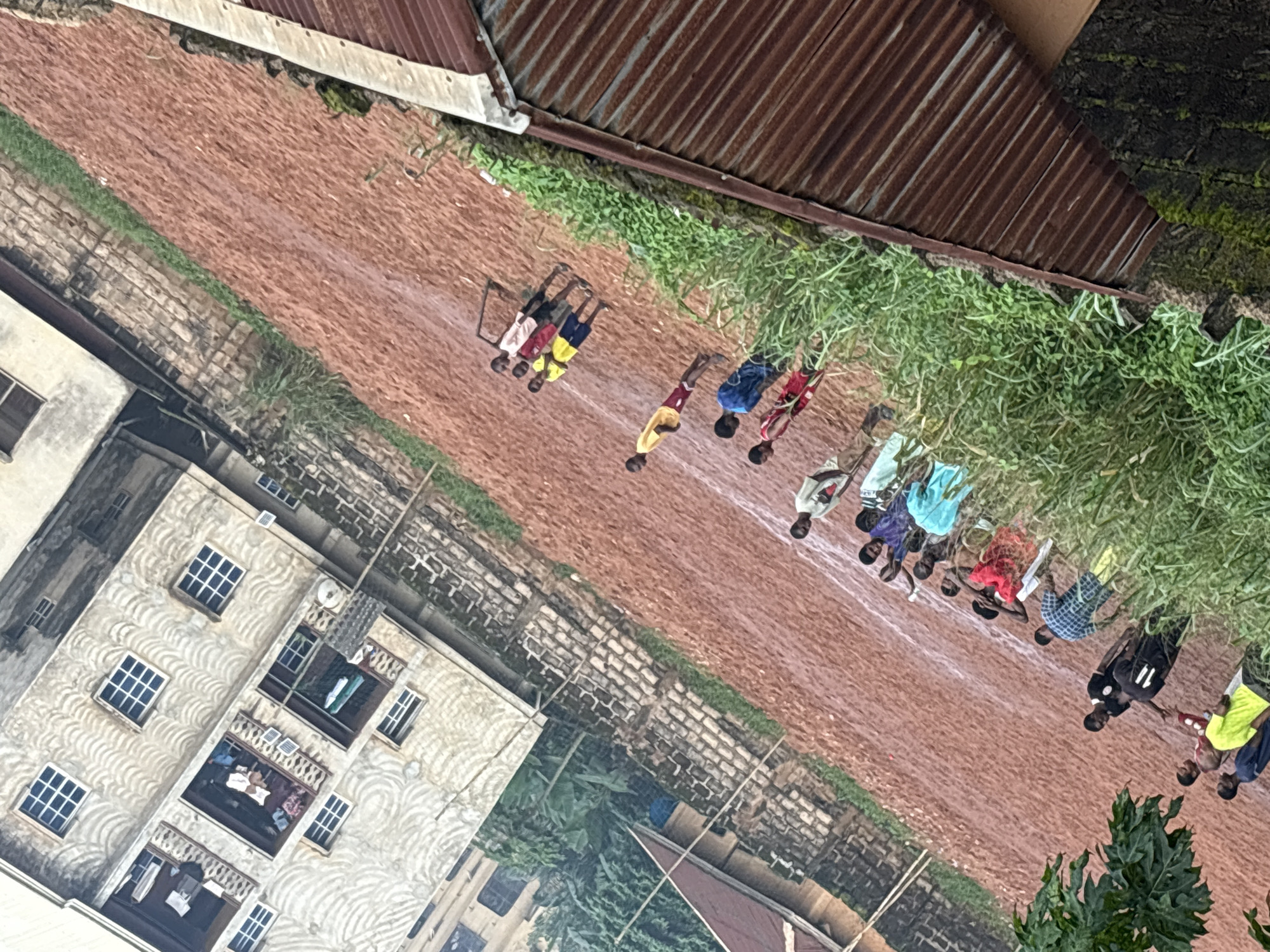 The image depicts a group of young people on a dirt field, possibly in Onitsha, Nigeria. The field appears to be a makeshift sports ground, with some faint white lines marking boundaries. In the background, a multi-story building with several windows and balconies is visible, suggesting an urban or semi-urban setting. Some of the people are standing in groups, while others are walking or interacting. The overall atmosphere appears casual, with the individuals dressed in various clothing. The ground is a reddish-brown earth, indicating it could be a common playing surface in the region. The presence of green foliage suggests it is likely daytime and potentially during a wetter season. The scene is shot from an elevated angle, looking down at the field and the people.