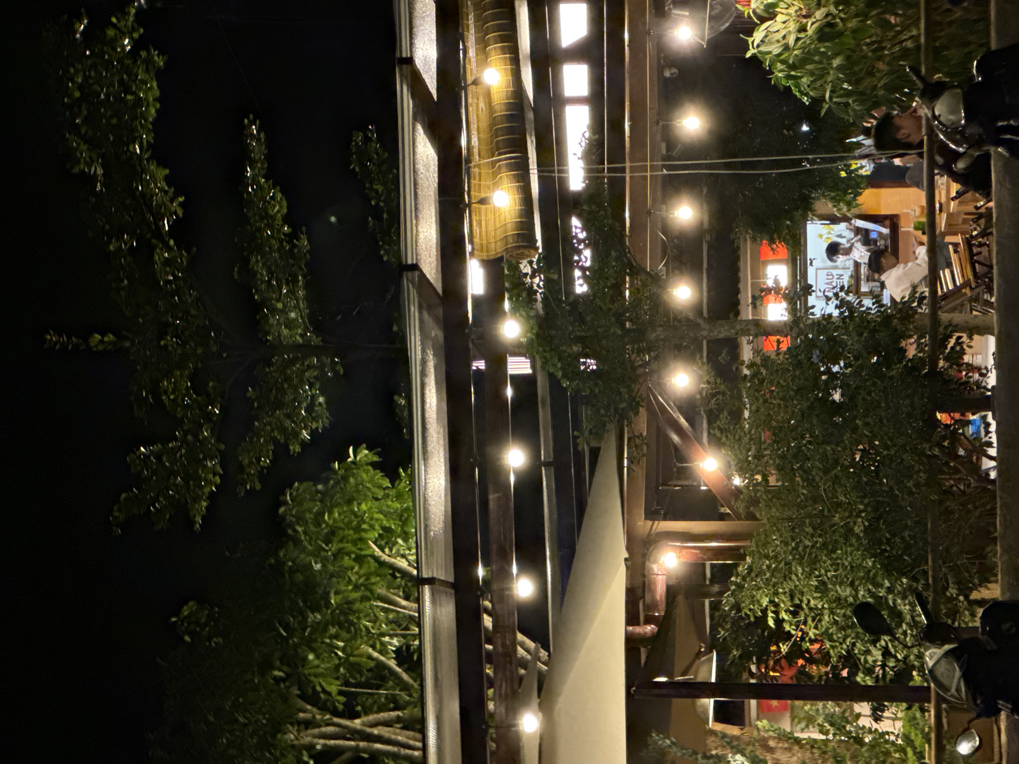 The image depicts a nighttime scene at an outdoor cafe in Lâm Viên - Đà Lạt, Vietnam. The cafe is illuminated by numerous string lights and overhead bulbs, casting a warm glow on the wooden structures and surrounding greenery. Several trees with dense foliage are visible, some reaching above the cafe's awning.
In the background, inside the cafe, two individuals are visible. One is wearing a white shirt and appears to be interacting with another person. A sign in the background is partially obscured but includes Vietnamese text.
In the foreground, a portion of a motorcycle is visible on the left side of the image, and another motorcycle is partially visible on the right. The overall atmosphere suggests a relaxed, casual setting for dining or socializing.