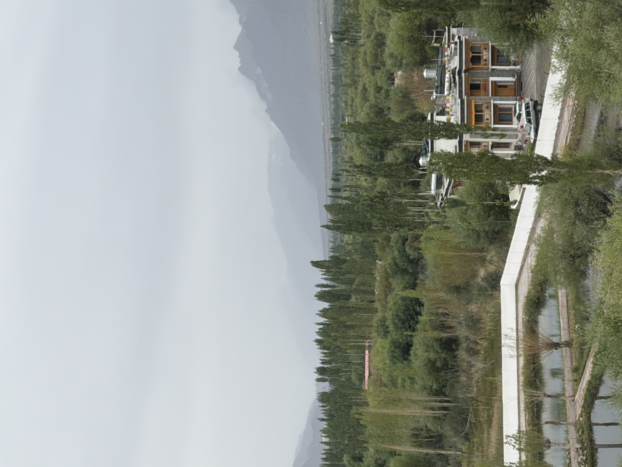 The image shows a view of the town of Thiksey, India, on an overcast day. In the foreground, there is a modern building with a white wall surrounding it. Beyond the wall, a body of water is visible, with trees and shrubs growing around it. Further in the background, a dense forest of tall, slender trees stretches across the landscape. A building with a traditional design, featuring multiple balconies and colorful prayer flags, is situated among the trees. Two vehicles, a white SUV and a darker vehicle, are parked in front of this building. The background is dominated by the silhouettes of mountains shrouded in mist, creating a hazy and atmospheric backdrop. The overall impression is one of a serene and somewhat subdued natural environment with touches of human habitation.