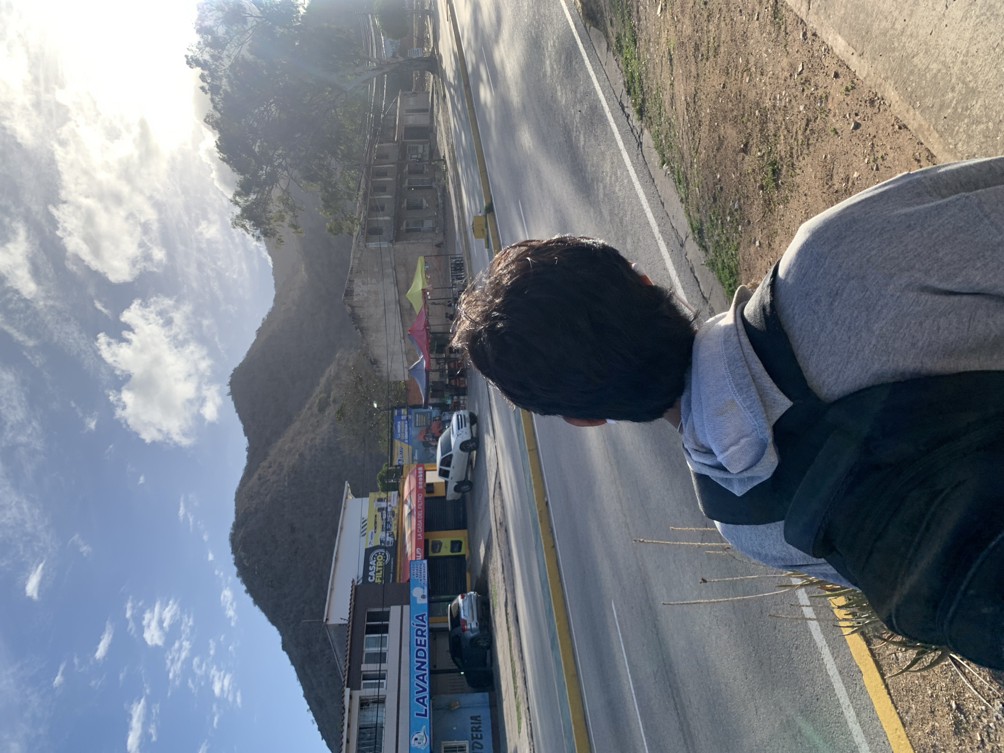 In El Valle del Espíritu Santo, Venezuela, a person wearing a grey hoodie and a black backpack stands with their back to the camera, looking towards a road and a mountainous landscape. The sun is bright, casting shadows and creating a glare in the sky. Across the road are several businesses, including "Lavandería" and "Casa del Filtro," with awnings set up outside. Buildings line the street, some appearing older and in disrepair, while others are more modern. The area is characterized by a dry, dusty environment with sparse vegetation and a clear, blue sky with scattered clouds.