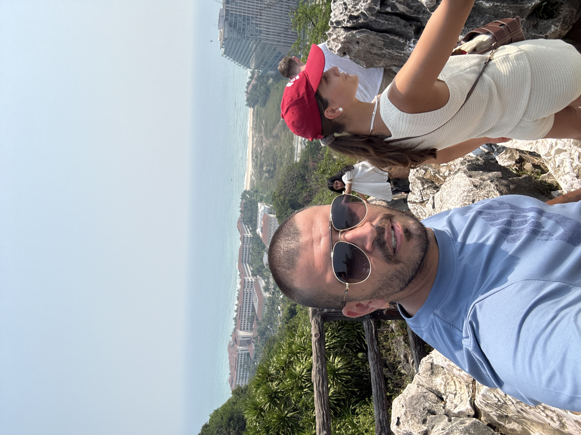 A daytime, medium shot captures a man in a blue t-shirt and sunglasses, taking a selfie. Behind him and slightly above, a young woman in a white outfit and red cap looks towards the coastline. Another person in a white shirt is visible in the background, along with a woman in a white dress. The scene overlooks a coastal city, likely Đà Nẵng, Vietnam, given the presence of beachside buildings and a hazy sky. The foreground consists of rocky terrain and some vegetation.