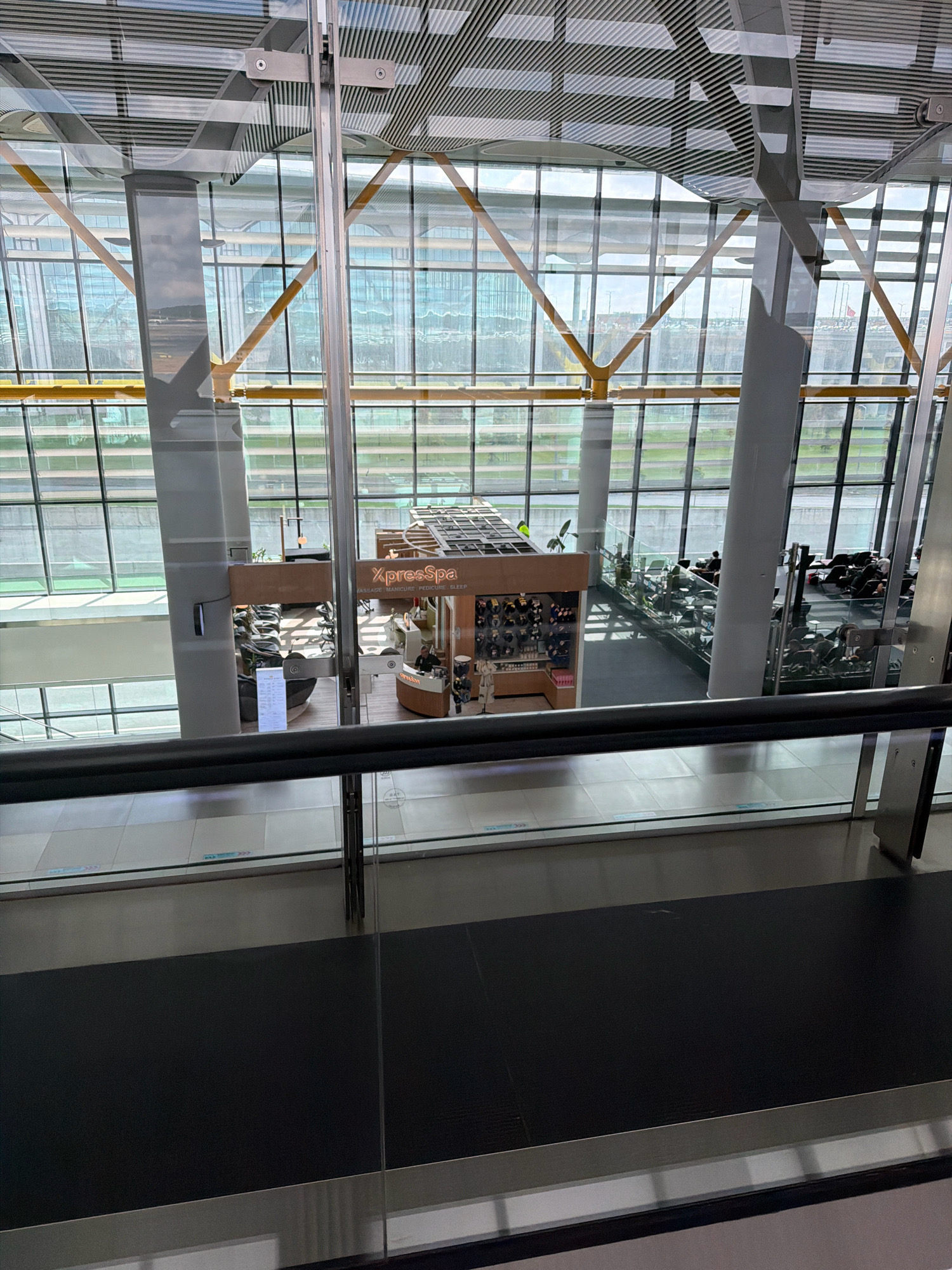 This image, taken from an elevated perspective, depicts the interior of a modern airport terminal in Arnavutköy, Turkey. Large glass windows dominate the scene, offering views of the exterior which includes tarmac, greenery, and distant structures. Inside, the architecture is characterized by a high ceiling with a grid-like metallic structure and exposed beams. Several large, white cylindrical pillars support the ceiling.
In the foreground, a dark, reflective railing runs horizontally across the frame, indicating a walkway or viewing platform. Below this railing, a dark, rectangular surface, possibly a carpet or floor covering, is visible.
Further into the terminal, a spa area labeled "XpressSpa" is situated. This spa features a curved counter where a person in a uniform is visible, seemingly interacting with a customer or employee. There are also several manicure stations with chairs and a display shelf containing various products. Adjacent to the spa, a waiting area is populated with individuals seated in chairs, some engaged with their devices. Other areas of the terminal show additional seating arrangements and what appear to be retail kiosks. The overall environment suggests a busy, functional space designed for travelers.