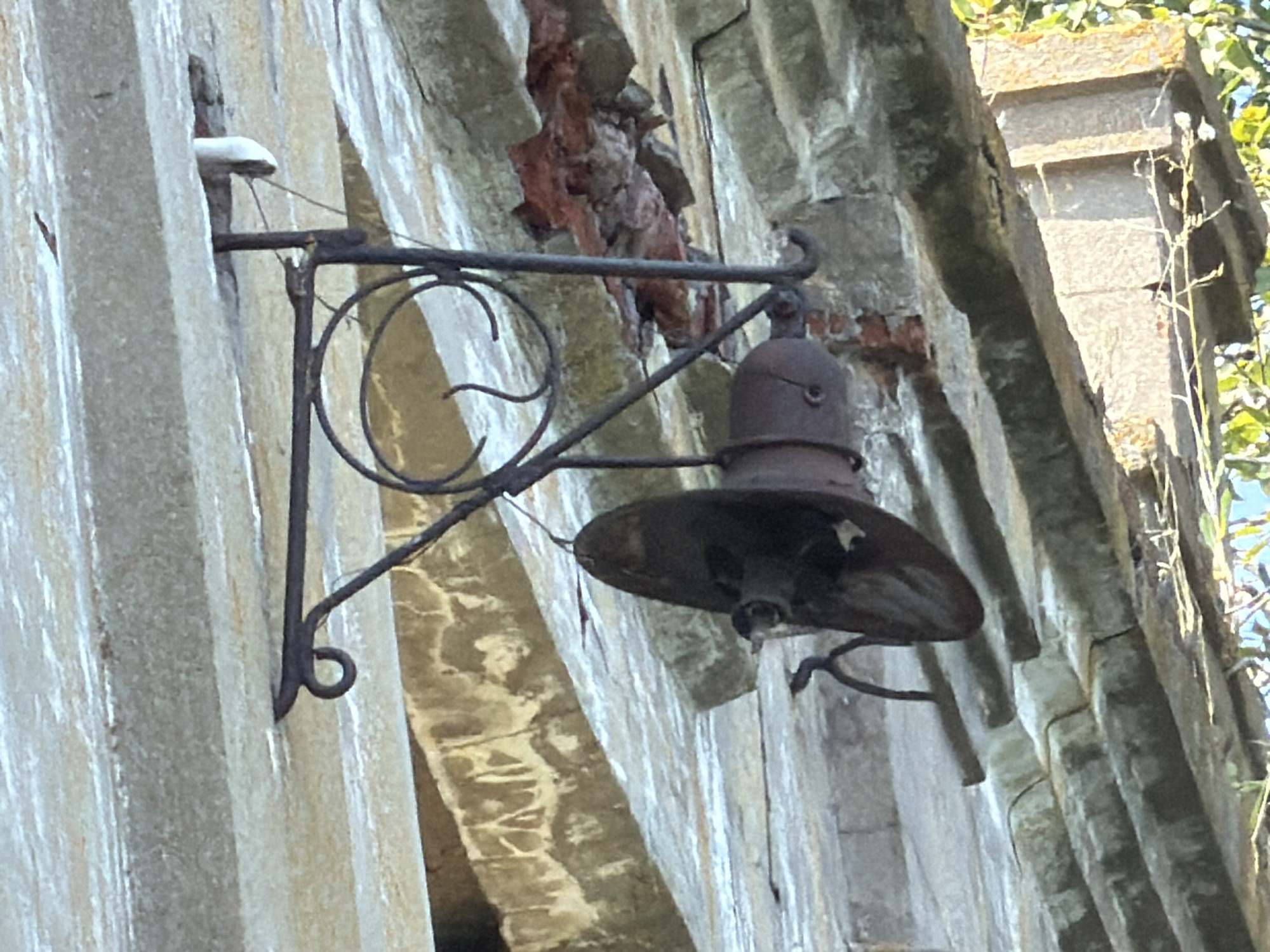 The image shows an outdoor scene depicting an old, rustic light fixture attached to a weathered, concrete or stone building. The fixture itself is made of dark metal and appears to be a lantern-style lamp with a bell-shaped top and a wide, downward-facing shade. The metal shows signs of rust and age. The building's surface is textured and appears to be composed of vertical, possibly precast concrete sections, with areas of damage and exposed reddish material, suggesting deterioration. The background shows some greenery, indicating an exterior setting, and the sunlight suggests it is daytime. The overall impression is one of disrepair and abandonment. The location in the city of Troekurovo, Russia is not visually evident from the image itself.