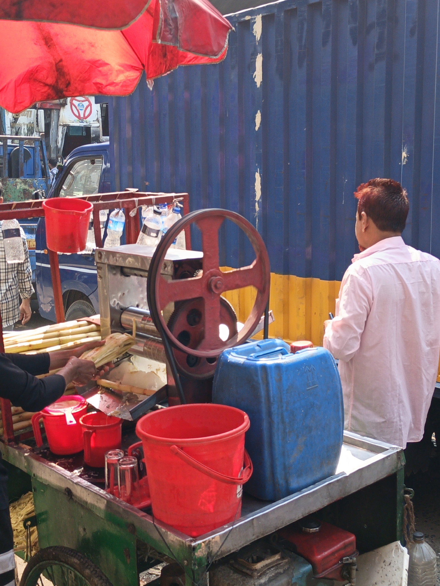 A street vendor operates a sugarcane juice cart in Dhaka, Bangladesh. The cart, under a red umbrella, has a sugarcane press with a large red wheel. A man in a light pink shirt stands on the right, facing the cart. Another man in a plaid shirt stands on the left side of the image, watching the sugarcane being fed into the press. The cart also has several red containers, a blue jerrycan, and bottles of water. A truck with blue corrugated siding is in the background.
