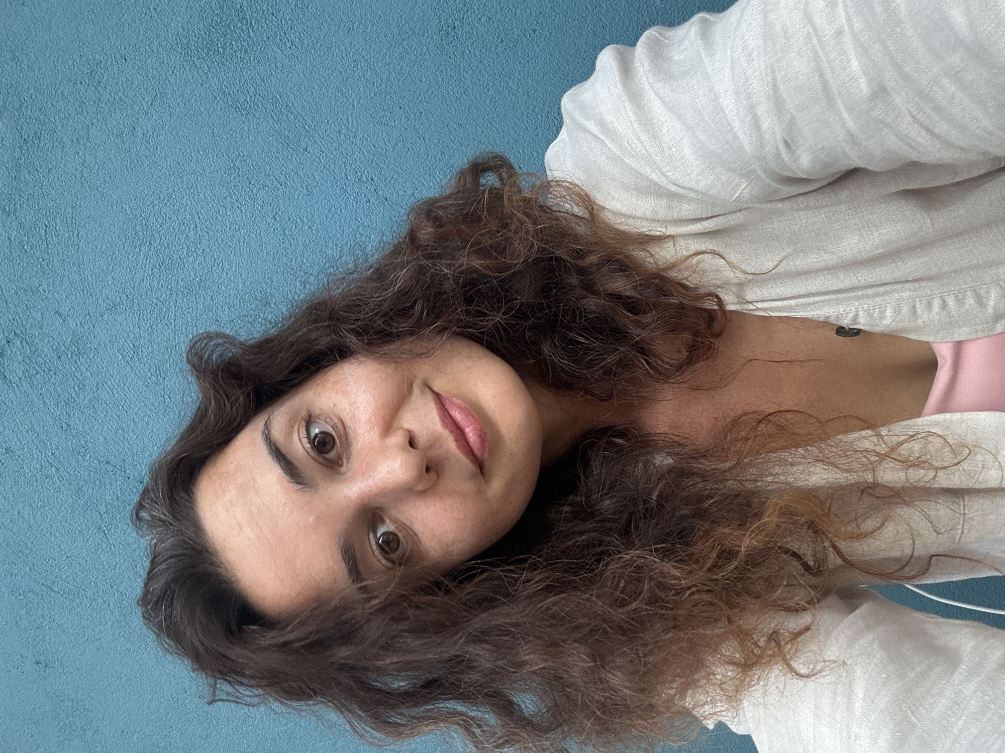 A selfie of a young woman with wavy brown hair, looking towards the camera with a slight smile. She is wearing a light-colored, collared shirt over a pink top. The background is a textured blue wall. The image appears to be taken outdoors. The scene does not contain any identifiable landmarks or elements that would suggest a specific location in Hoi An, Vietnam.
