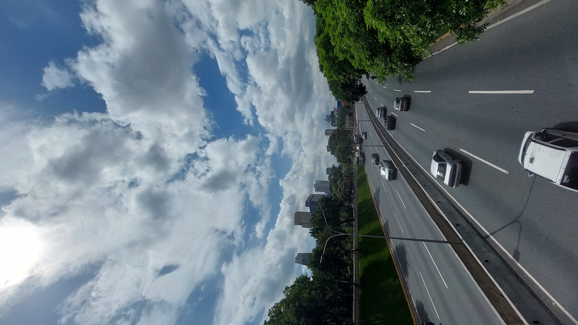 The image shows a view of a highway in Caracas, Venezuela, likely taken from an elevated position. The highway has multiple lanes with cars moving in the same direction. A median separates the highway from a green space with trees on either side. The sky is blue with scattered white clouds. A few buildings are visible in the background, suggesting a city environment.