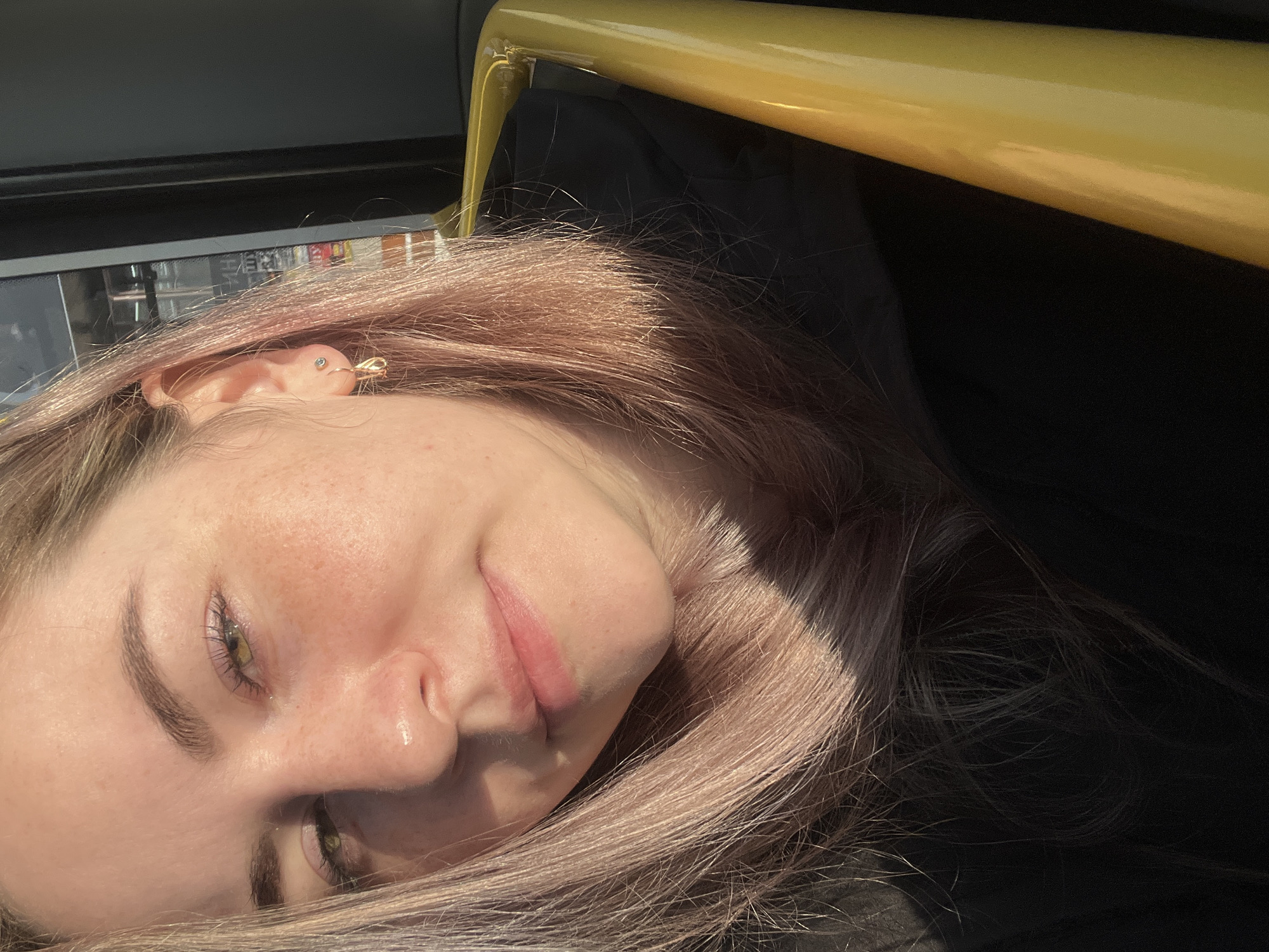 A young adult female is depicted in a medium-shot portrait inside a vehicle, located in Chaikovsky, Russia. She has fair skin, prominent freckles on her nose and cheeks, and green eyes directed towards the viewer. Her hair is long, straight, and light brown with a pinkish tint, illuminated by direct sunlight on the left side of her head. The subject wears a dark-colored jacket or top. Her right earlobe is adorned with two earrings: a small stud in the upper lobe and a larger, gold-colored teardrop-shaped hoop in the lower lobe. Strong sunlight casts a bright highlight across her nose and left cheek, creating contrast with shadowed areas. A distinct, glossy yellow grab rail or bar from the vehicle's interior is visible along the right side of the frame. The background consists of blurry interior elements or reflections.
