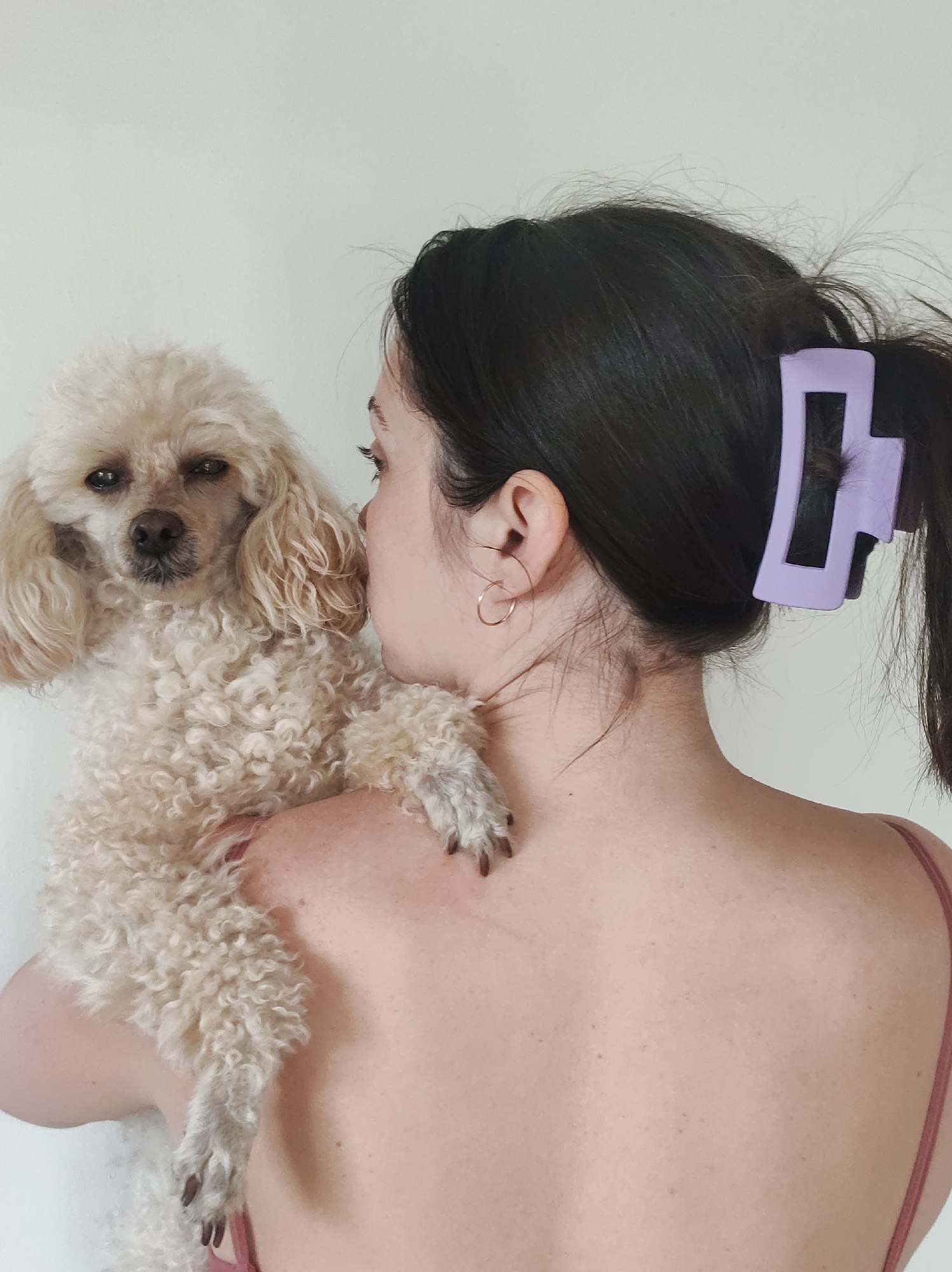 A woman with dark hair pulled back in a ponytail, secured with a lavender claw clip, is holding a cream-colored poodle. She is wearing small gold hoop earrings and a thin-strapped top. The background is a plain white wall. The location is unknown.
