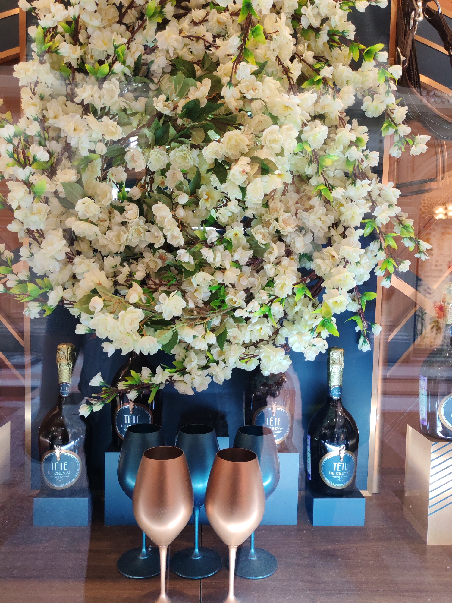 This media file captures a still life arrangement, likely a product display or decorative scene. The foreground is dominated by a large, lush arrangement of white faux flowers, possibly cherry blossoms, cascading from the top of the frame. Beneath the floral display, two bottles of what appears to be champagne or sparkling wine are prominently featured. Both bottles bear labels with the text "TÊTE DE CHEVAL" and "MÉTHODE CLASSIQUE BRUT," suggesting they are the same brand and type of beverage. The bottles are presented on dark, rectangular pedestals.
Flanking the bottles are four wine glasses. Two of these glasses have a dark, metallic finish, possibly black or dark blue, with tall stems and wide bowls. The other two glasses have a striking copper or rose gold finish, also with slender stems and flared bowls. These glasses are arranged in a way that suggests a serving or tasting setup.
The background is dark and somewhat out of focus, with hints of warm lighting and geometric decorative elements, suggesting an indoor setting such as a store window display, a bar, or a dining area. The overall impression is one of elegance and celebration, with the floral abundance and sparkling wine bottles evoking a sense of luxury or festivity. There are no people visible in the image. The lighting appears to be artificial, creating a slightly reflective quality on the glass and metallic surfaces. The image seems to be taken from a slightly elevated perspective, looking down on the arrangement.