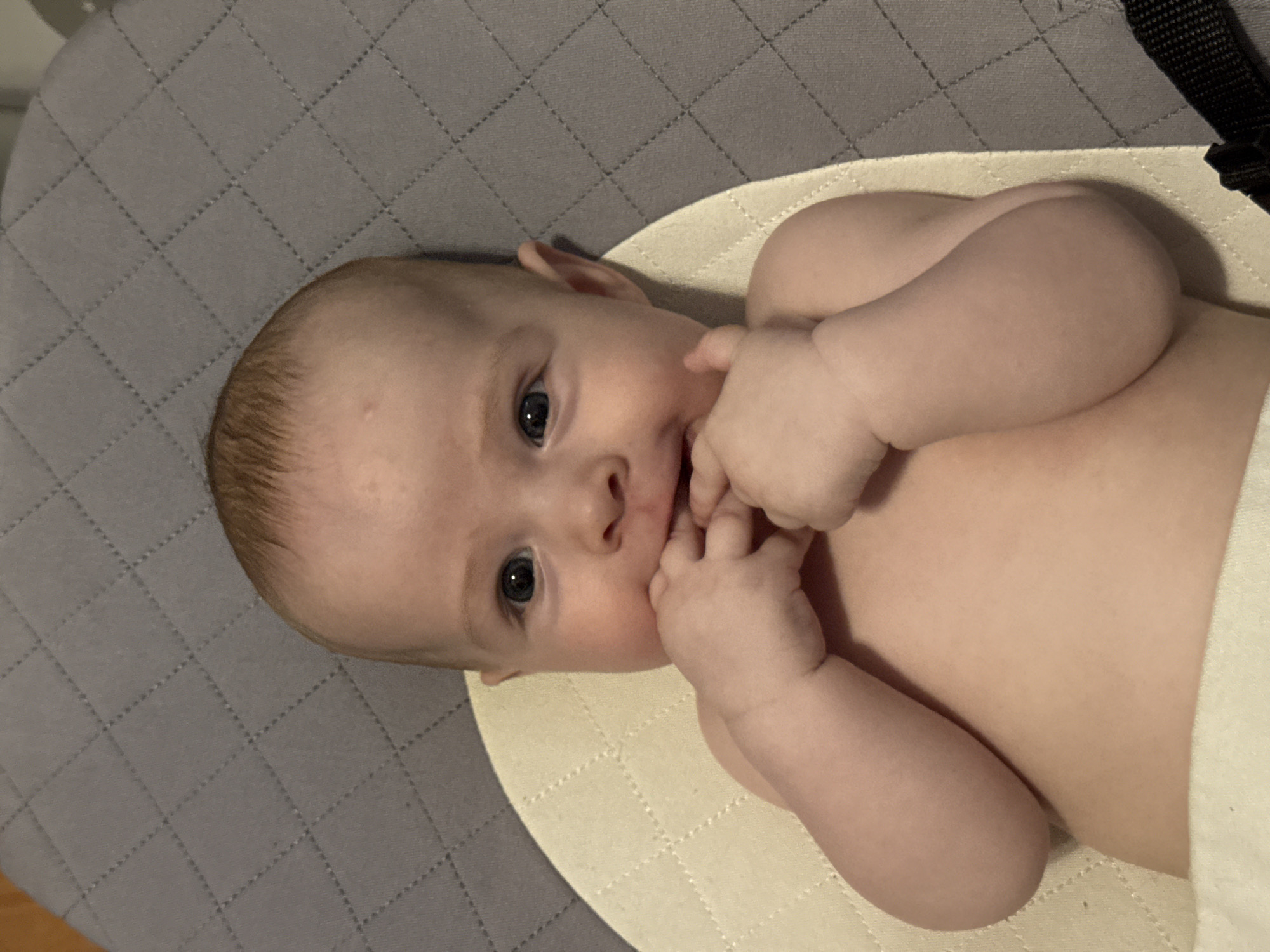 A baby lies on a quilted, off-white surface with a gray quilted pad in the background. The baby is fair-skinned, with light hair and blue eyes. The baby has fingers in its mouth and is looking at the camera. Part of a black strap is visible at the upper right of the image. There are no indications of the location being Chaikovsky, Russia.