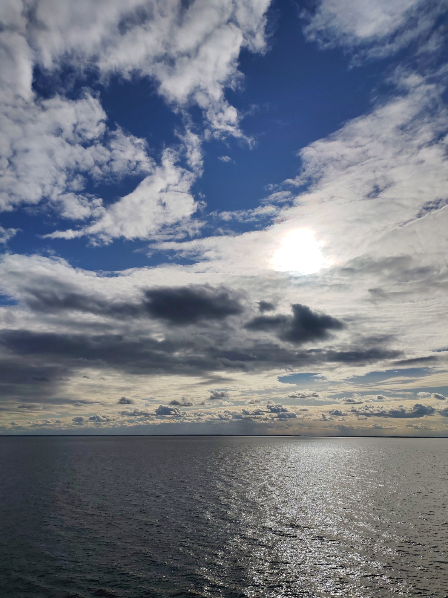 This vertical shot captures a vast and dynamic seascape or large lake view, likely from Prislon', Russia. The image is divided between an expansive body of water in the foreground and midground, and a dramatic sky above, separated by a distant, low horizon line.
The sky is a prominent feature, displaying a complex mix of cloud formations and clear blue patches. In the upper regions, particularly the upper left, sections of deep blue sky are visible amidst scattered white, cumulus-like clouds. Below this, a dense band of darker grey clouds stretches horizontally, creating a sense of depth and varying light conditions. The sun, a brilliant white orb, is partially obscured by these clouds on the upper right side, casting an intense, luminous glow. This indicates a daytime setting, likely late morning or afternoon.
The large body of water beneath the sky appears dark greyish-blue, with ripples and small waves visible on its surface. A distinct, shimmering path of sunlight reflects brightly on the water, extending from the horizon towards the bottom right of the image, directly reflecting the obscured sun. The horizon itself is a faint, dark line, suggesting either a very flat, distant shoreline or an open expanse of water.
The scene conveys a sense of natural grandeur and tranquility. The weather appears partly cloudy, with both bright sunlight and dark cloud cover contributing to a dynamic visual atmosphere. There are no visible people, man-made objects, or text within the frame, emphasizing the natural landscape.