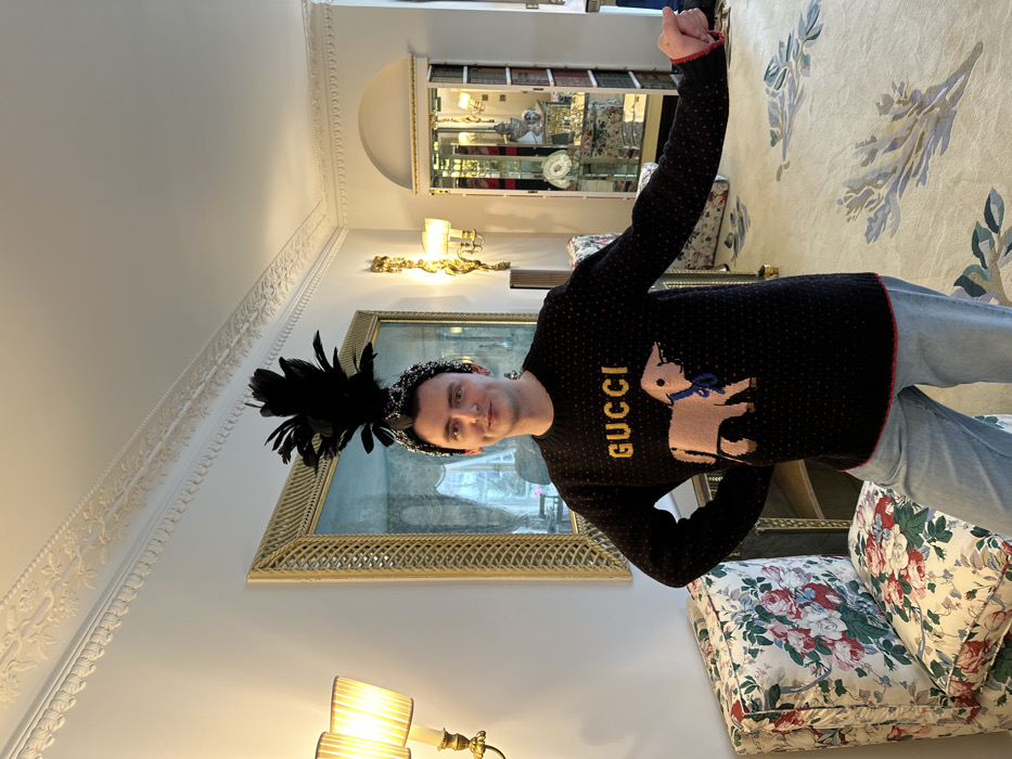 A single adult male stands centrally within a luxuriously decorated room in London, United Kingdom. He wears a black long-sleeved sweater adorned with "GUCCI" in yellow text and a light brown lamb graphic featuring a blue bow, paired with light blue jeans. A black feather plume is affixed to a sparkling beaded headband on his head. The subject faces forward, exhibiting a subtle smile, with his left hand resting on his hip and his right arm extended slightly outwards.
The room features off-white walls with elaborate white plaster coving and ceiling moldings. To the left, a floral-patterned armchair is partially visible, adjacent to a gold-toned wall sconce with two shaded lamps emitting warm light. A large mirror with a wide, ornate gold-textured frame occupies the background, reflecting an interior space or outdoor view. To the right, another identical wall sconce is present, alongside a tall glass display cabinet containing decorative porcelain and glass items. The floor is covered by a light-colored carpet featuring a large, stylized botanical pattern in shades of blue, green, and cream.