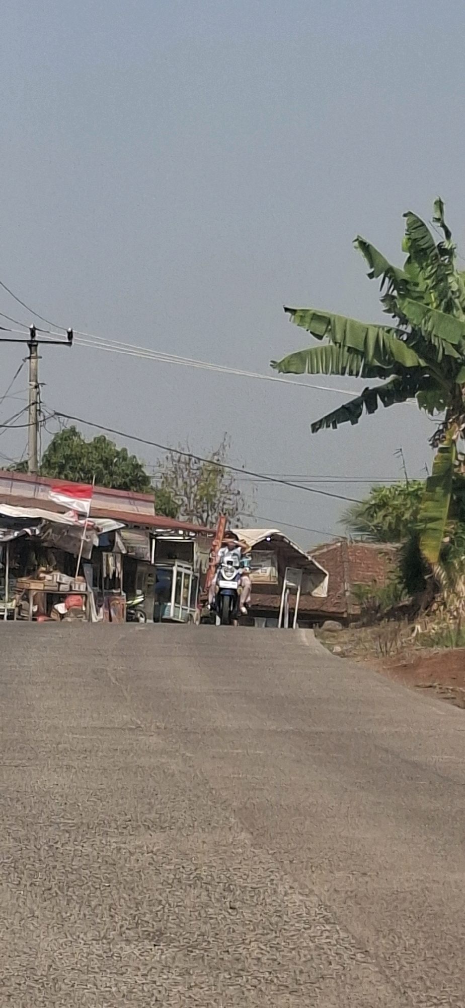 A person rides a motorcycle up a paved road lined with small roadside structures. The person is wearing a white shirt and dark pants. The motorcycle is dark blue with a white seat. The structures appear to be shops or stalls. One stall has a red and white flag displayed. The background features trees and a hazy sky. The scene is consistent with a rural or semi-urban area in Cibinong Hilir, Indonesia.