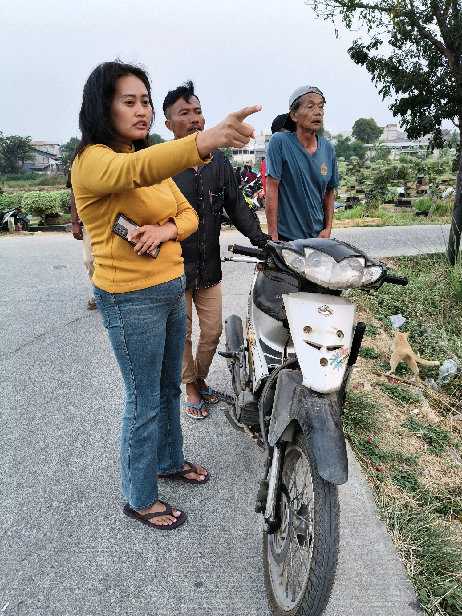 In Mangunjaya, Indonesia, three individuals stand near a parked motorcycle on a paved surface. A woman in a yellow long-sleeved shirt and blue jeans holds a smartphone in her right hand and points with her left hand towards the left of the frame. Beside her, a man in a black shirt and brown pants looks in the same direction. To their right, an older man in a blue t-shirt and a grey cap gazes forward. A ginger cat is visible in the grassy area to the right of the motorcycle. The background features buildings and trees under an overcast sky.