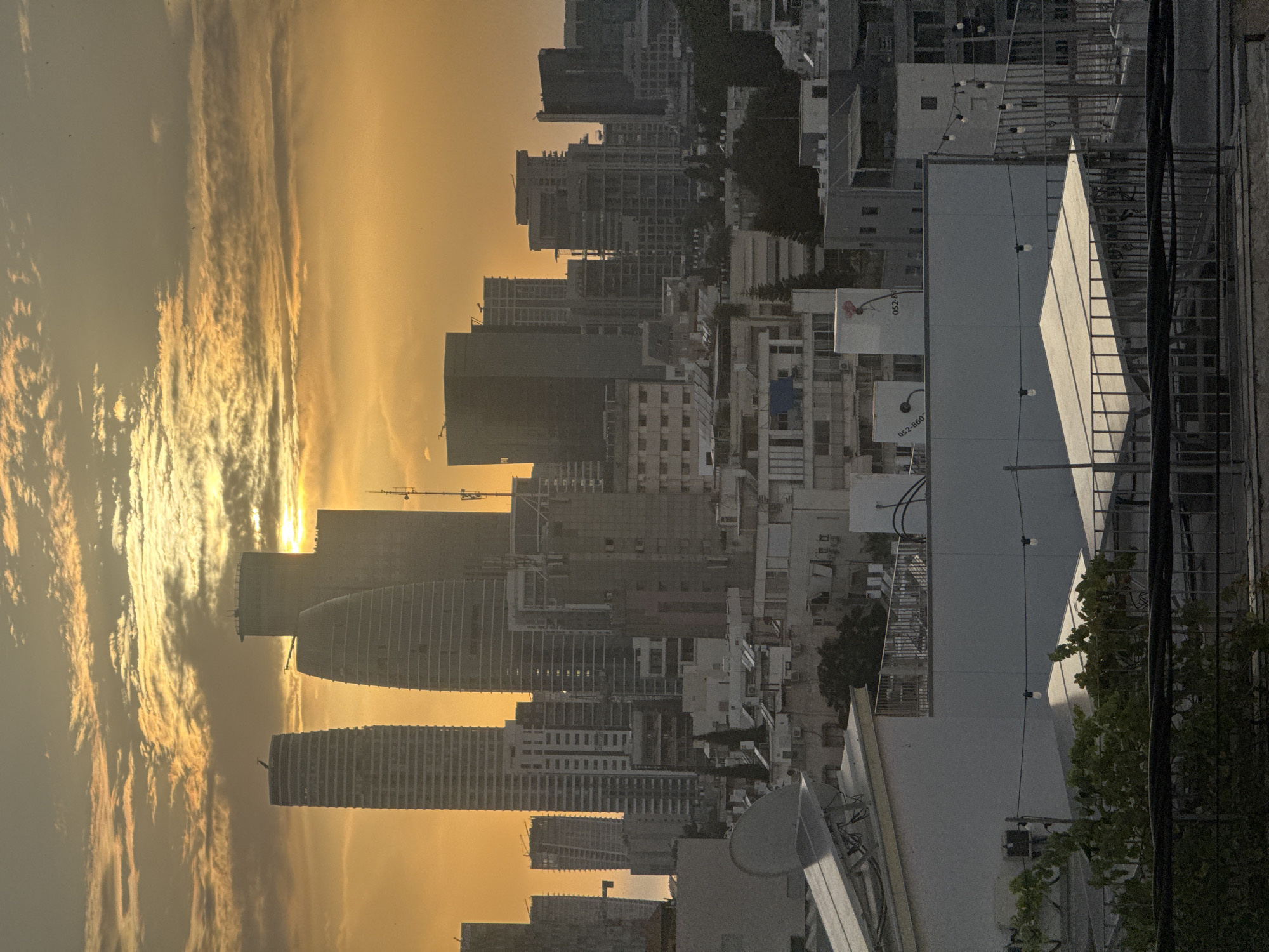 The image displays an urban skyline in Ramat Gan, Israel, captured during either sunrise or sunset, evidenced by the sky's striking golden-orange and yellow tones. The sun, a bright point, is partially visible near the horizon, casting a warm, dramatic light on elongated, streaky clouds.
The scene is characterized by numerous modern high-rise buildings, including skyscrapers and apartment blocks of varying heights and architectural styles. Several structures appear to be under construction, with visible concrete frameworks and at least one construction crane, highlighting ongoing urban development. Many distant buildings are silhouetted against the brightly lit sky.
In the foreground and mid-ground, rooftops feature typical urban fixtures such as air conditioning units and satellite dishes. A distinctive white building with a sloped roof and a string of decorative globe lights is prominent. Sparse greenery and trees punctuate the built environment.
Visible text on white signs with a red circular logo (resembling a stylized flower or cherry) includes partial phone numbers or codes, "052-860" and "052-86." No people are visible. The image conveys the atmosphere of a developing, modern city at a transitional time of day.