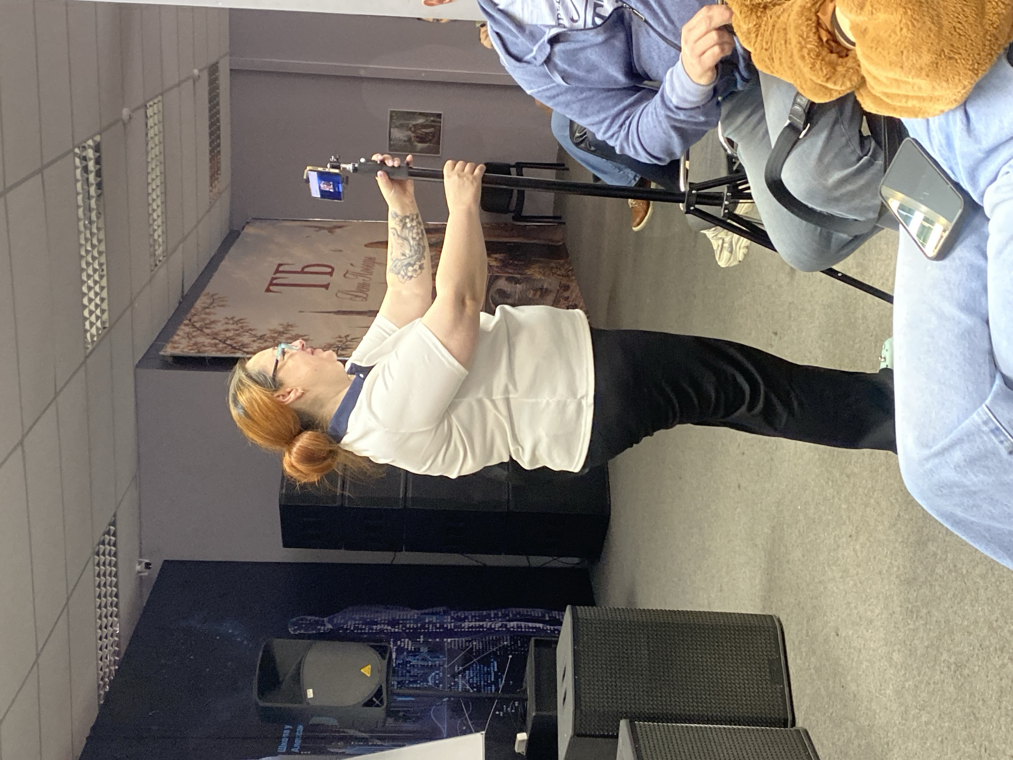 A person in a white shirt and black pants holds a black selfie stick with a phone attached at the end, possibly filming or taking a photo. The person is wearing glasses and has their red hair tied in a bun. Their arms are raised, holding the selfie stick above eye level. The scene appears to be indoors, possibly in an event hall or studio, with a large backdrop featuring city skylines and celestial designs. Audio equipment, including speakers, is visible in the foreground. The location is in the city of Unknown, Unknown.