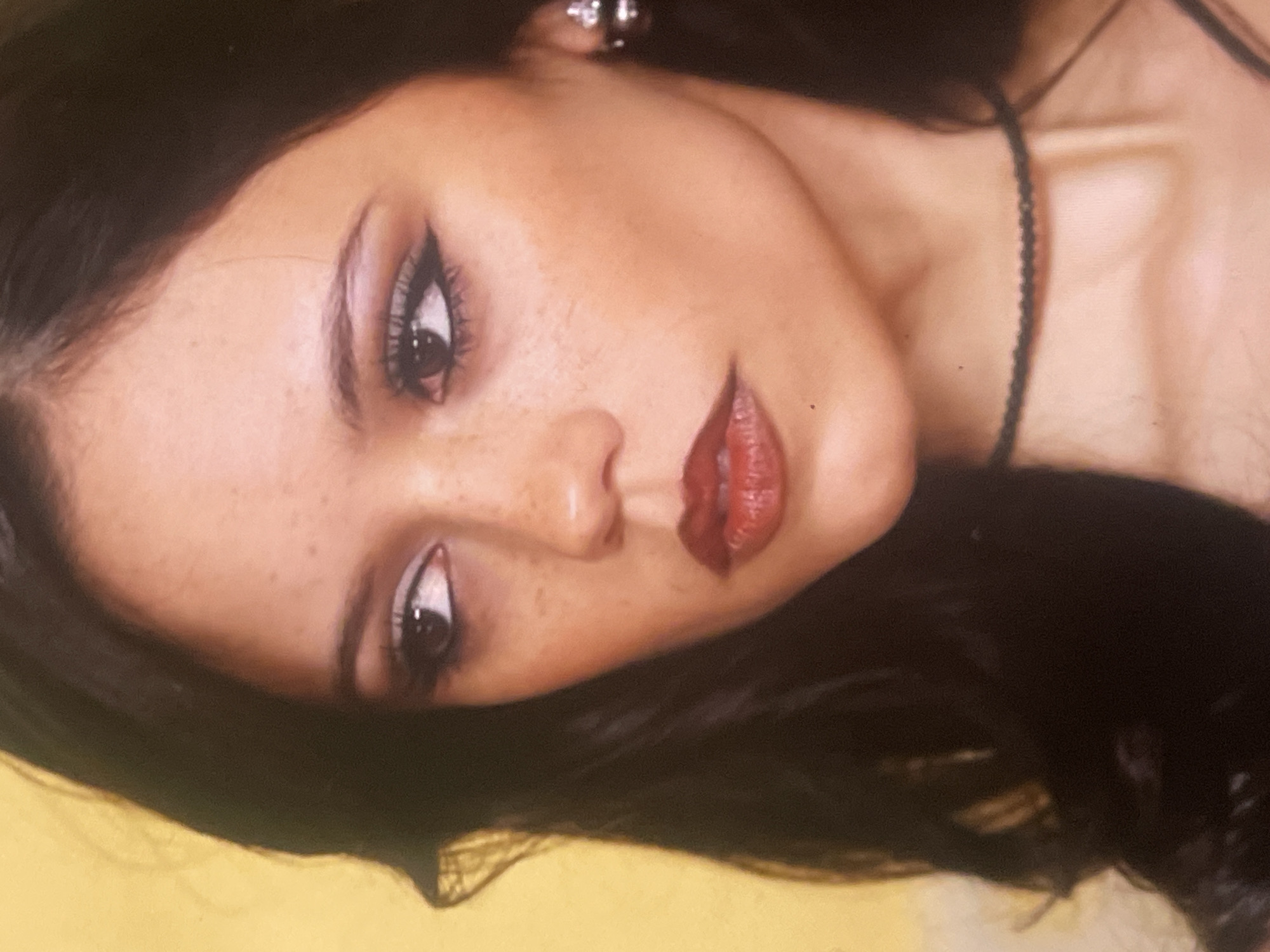 The image is a close-up portrait of a young woman, likely taken in Poltava, Ukraine, with her head tilted to the right. She is looking directly towards the viewer with a composed expression.
The subject has dark hair, which frames her face, and prominent dark eyes. Her eyes are accentuated with dramatic eyeliner, creating a winged look, and full, dark eyelashes. Her lips are full and glossy, colored with a deep red lipstick. Visible freckles are scattered across her nose and cheeks, complementing her warm skin tone. On her visible left ear, she wears a small, silver-toned earring. A thin, dark, possibly beaded necklace or strap of clothing is visible around her neck and extending towards her chest.
The background is a solid, bright yellow, suggesting a studio setting or a plain backdrop. The lighting appears soft and even, highlighting her facial features. There are no visible details regarding the time of day or weather. No text is visible in the image. The overall impression is that of a stylized and intimate portrait.