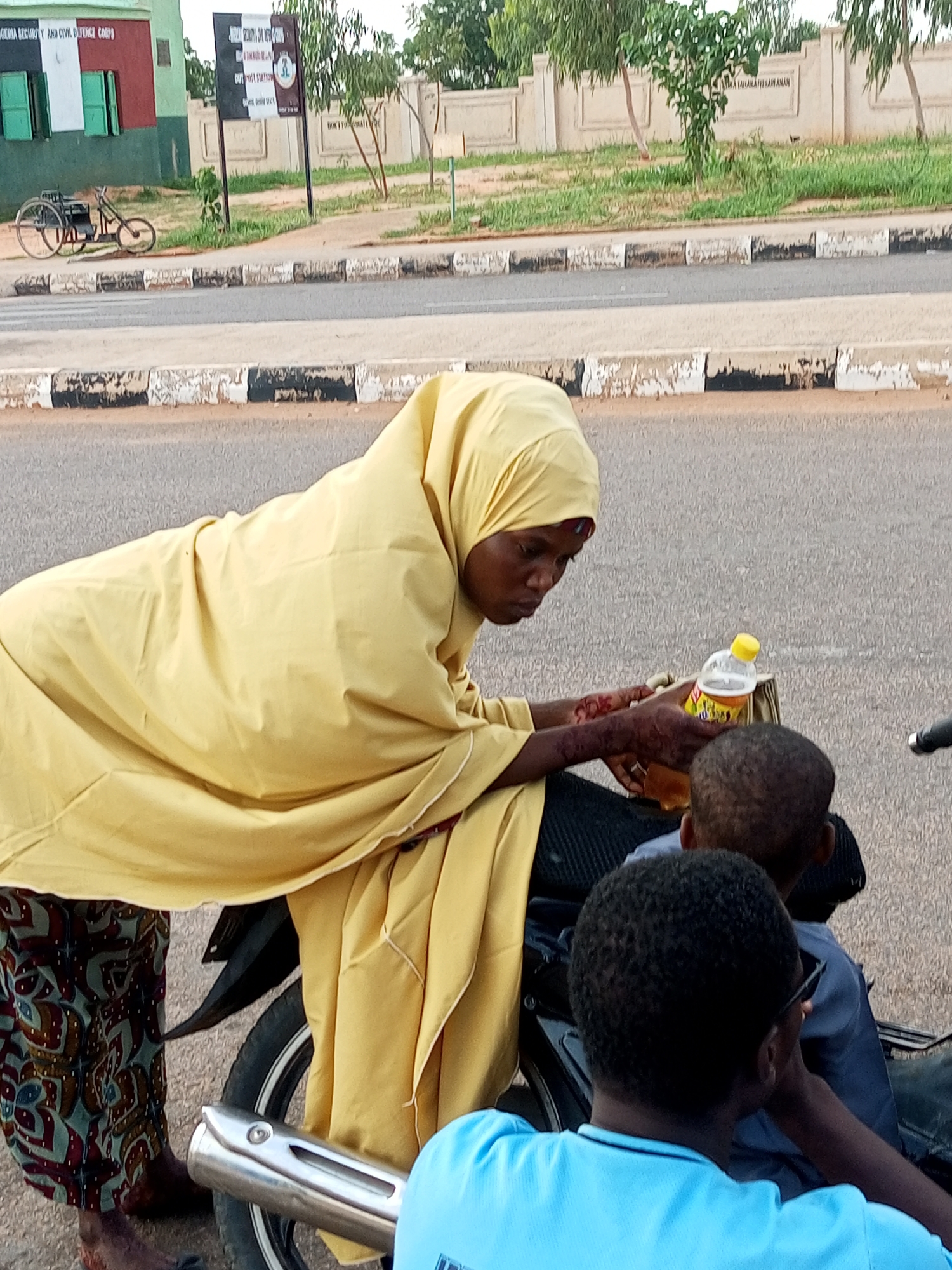 In an outdoor scene in an unknown city, a person wearing a yellow hijab and a patterned dress leans over a motorcycle. They are holding a yellow-capped bottle of liquid. Two children are seated on the motorcycle, facing away from the viewer. One child wears a blue shirt and sunglasses, while the other wears a dark shirt. The background shows a paved road with a concrete barrier, a sidewalk, and a building with signs. A bicycle is parked to the left of the building. The scene appears to be in a public area, possibly near an official building given the signage.