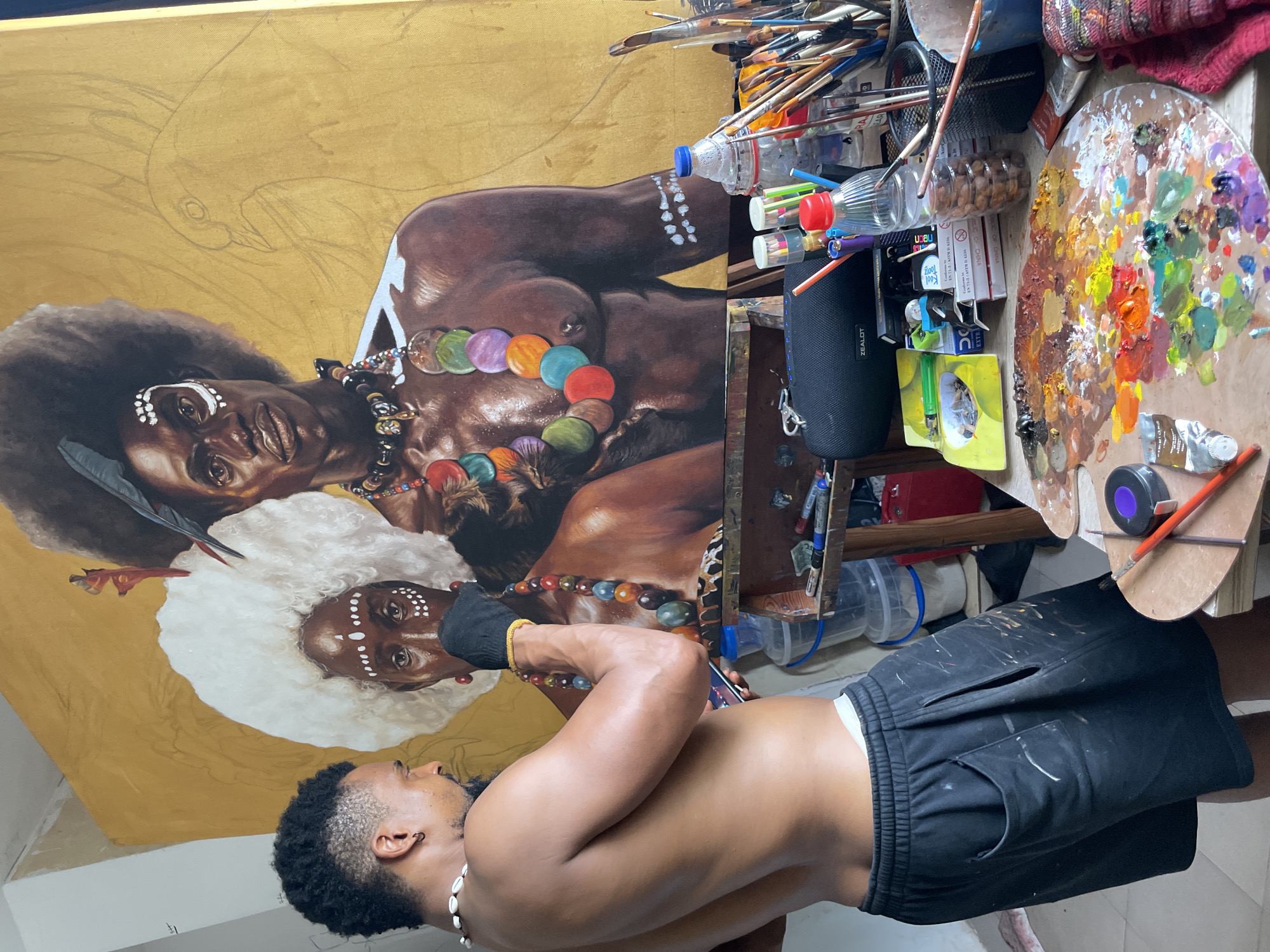 The image shows a shirtless dark-skinned male artist wearing black shorts with paint splotches, a shell necklace, and what appears to be a compression sleeve on his left hand, standing in front of a large painting of two dark-skinned women adorned with tribal jewelry and markings. The artist is holding a paint brush as if in the process of painting. The painting itself is mostly finished, with a golden background. The location appears to be an art studio with paint supplies such as brushes, bottles, containers, a Zealot-branded speaker, and a paint palette cluttered to the side. It is unclear if this is Ado, Nigeria.