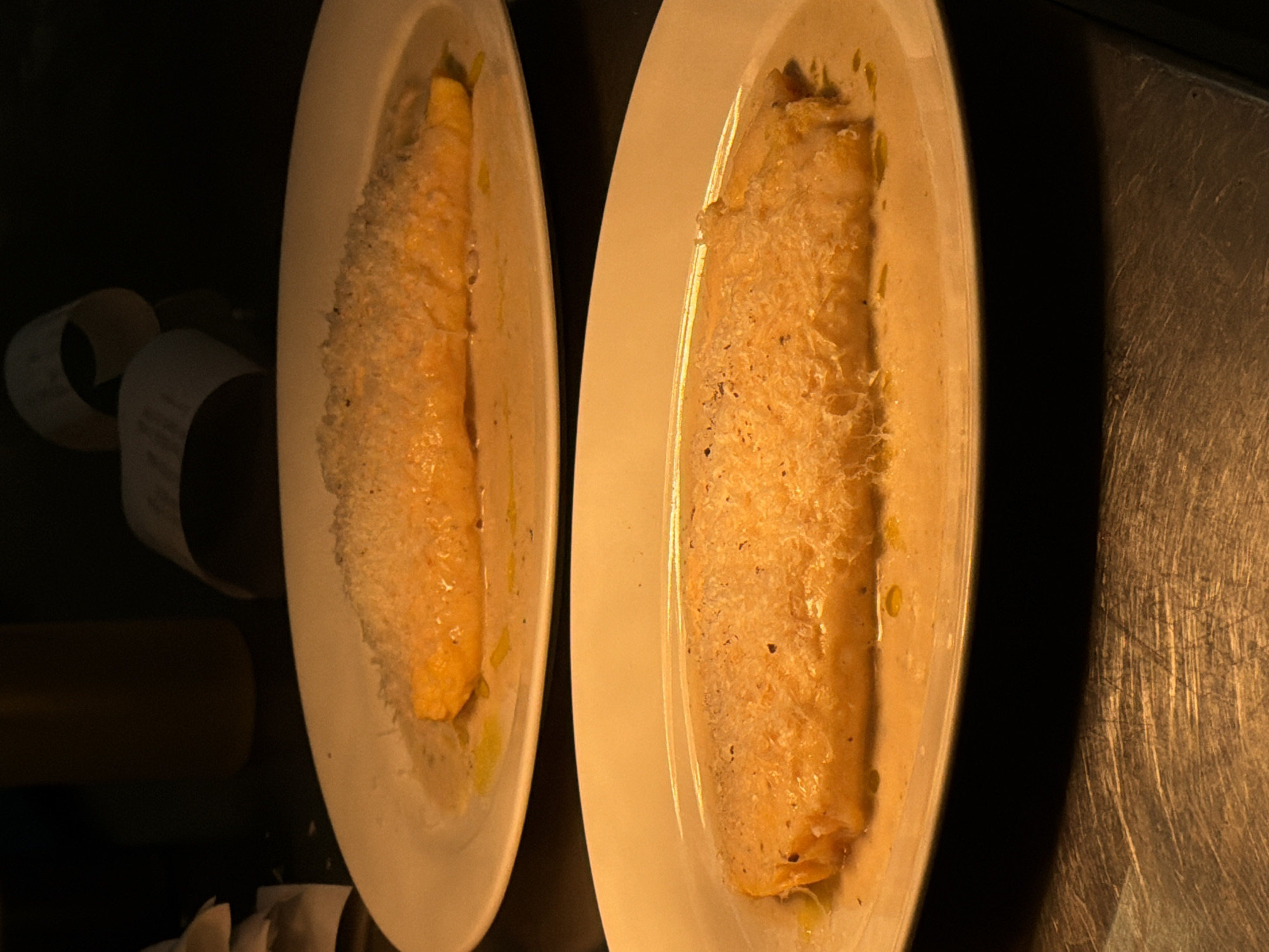 The image depicts two white, oval plates arranged side by side on a dark surface, possibly a table or countertop. Each plate contains a piece of fish, likely salmon, covered with a light-colored topping that appears to be grated cheese or breadcrumbs. The fish has a slightly glistening surface. The lighting is uneven, creating shadows and highlighting the textures of the food and surfaces. To the left of the plates, partial views of objects are visible, including what appears to be a roll of paper (possibly a receipt) and a cylindrical object, possibly a bottle. The surface to the right of the plates has a metallic appearance with visible scratches or wear. The location in Belgrade, Serbia, is not discernible from the visual information provided in the image. There are no people depicted.