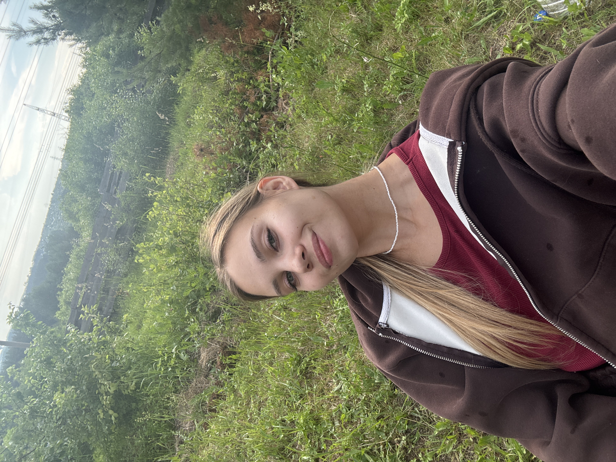 A young woman with blonde hair smiles at the camera in a selfie taken outdoors. She is wearing a dark brown zip-up hoodie over a maroon top, and a delicate white beaded necklace. The background is a lush, green grassy area with trees and some distant structures that appear to be industrial, possibly power lines and a crane, suggesting a location near an industrial zone or infrastructure in the city of Chaikovsky, Russia. The sky above is partially cloudy.