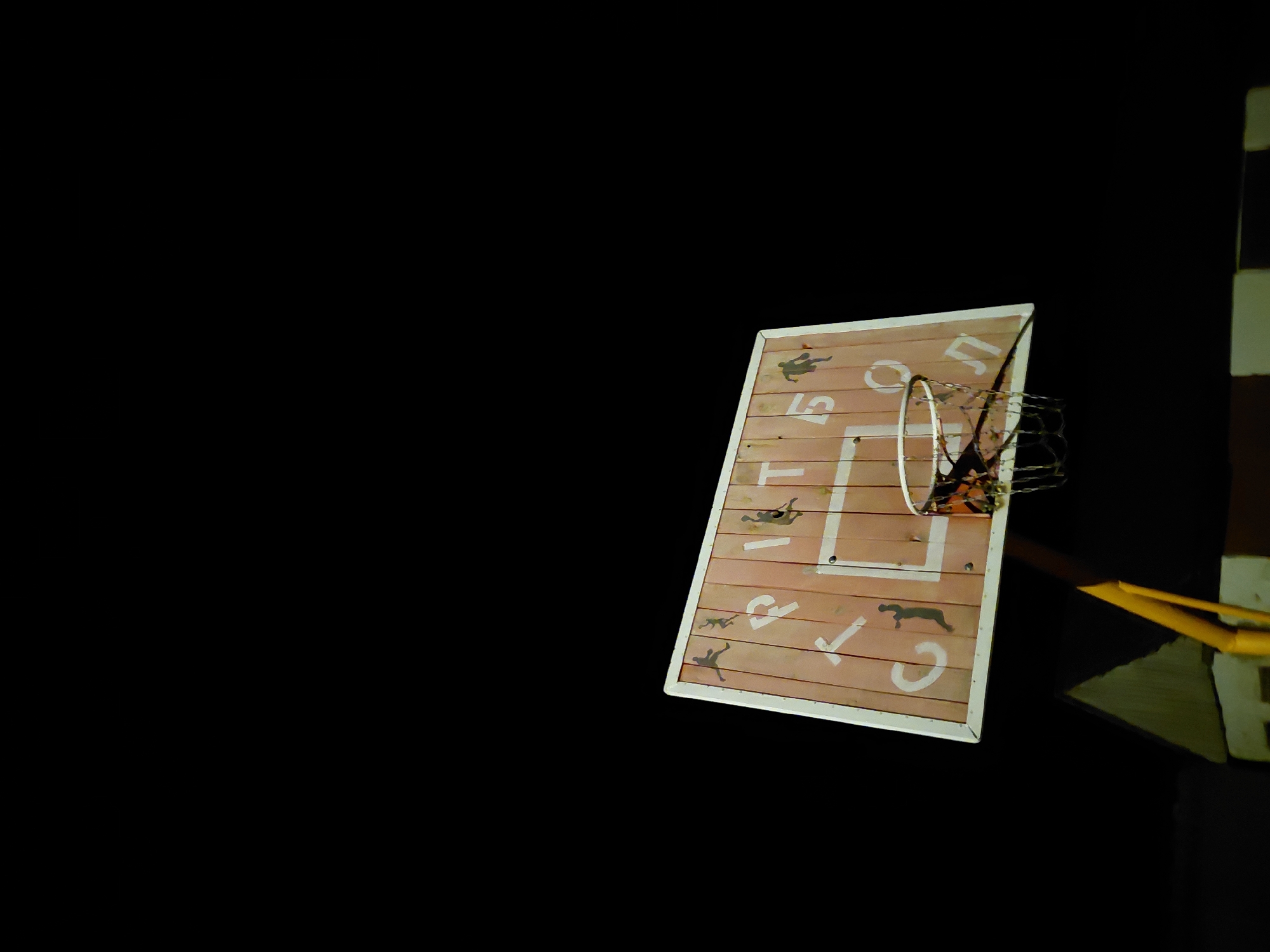 The image displays a basketball backboard and hoop against a completely black background, suggesting nighttime or a dark environment where only the subject is illuminated. The backboard is made of wooden planks and has a somewhat rustic appearance. It is adorned with white markings, including a rectangle and a square around the hoop's center.
A metal chain net is attached to the hoop. To the right of the backboard, a yellow structure, likely part of the support pole, is partially visible. The entire setup is seen from an angled, almost tilted, perspective.
Notable details on the backboard include white Cyrillic letters, which spell out "СПОРТ" (SPORT). The letters "С", "П", "О", "Р", and "Т" are clearly discernible, though some are partially obscured or cut off by the frame. Additionally, several dark, stylized silhouettes of people engaged in athletic activities, such as running or jumping, are painted onto the wooden surface of the backboard.
No activity is taking place; the image focuses solely on the basketball equipment. The lighting highlights the backboard and hoop, making them stand out against the dark void.