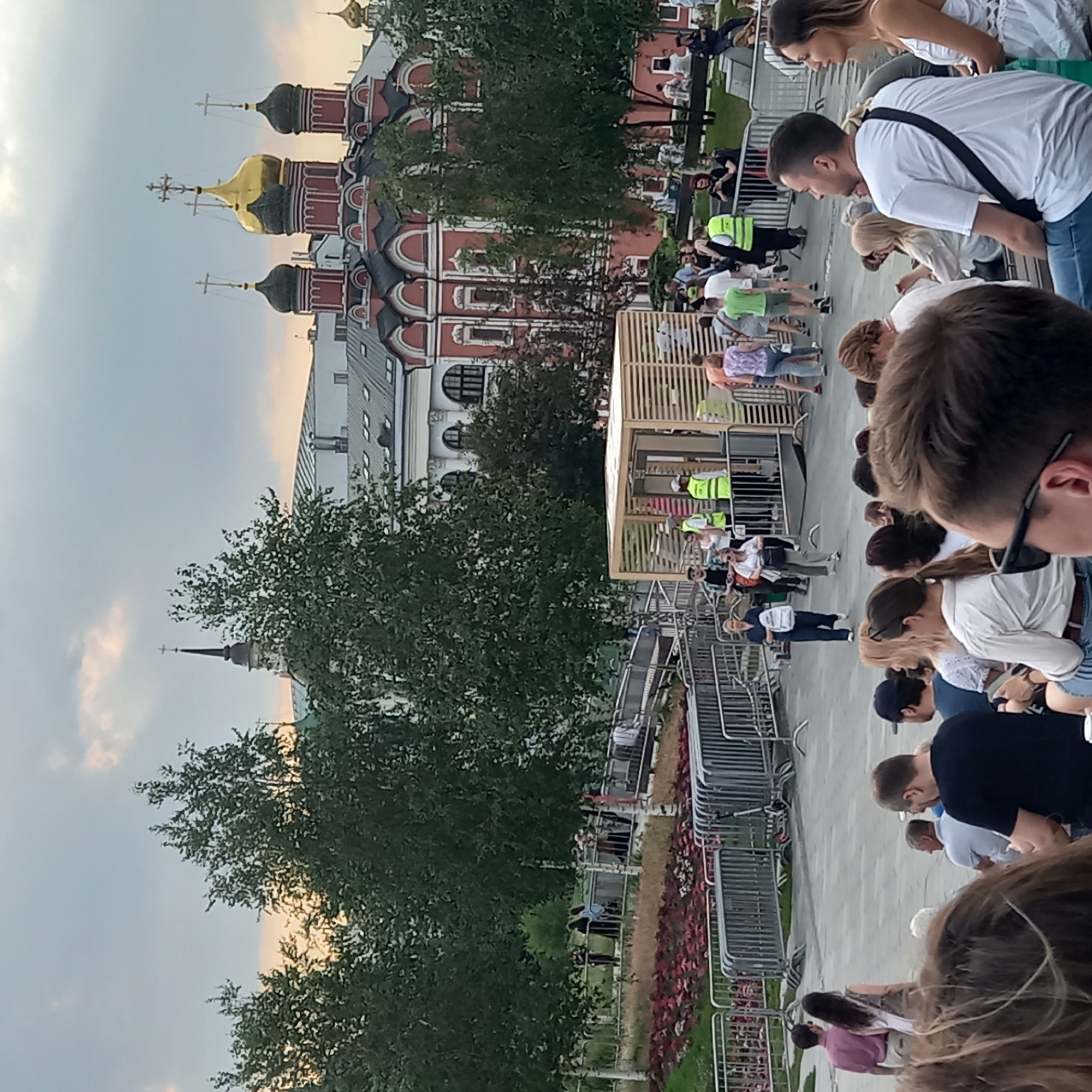 This image depicts an outdoor gathering in a public space, likely a park or plaza, with a church building in the background. The church features multiple domes, with one prominent golden dome and several smaller, darker domes. The building is ornate, with red walls and white decorative elements. The sky is overcast with hints of warmer light peeking through.
In the foreground and midground, numerous people are present, some standing and others seated. Many individuals appear to be spectators, facing towards an unseen point of interest or event. Several individuals in bright yellow vests are visible, suggesting they may be event staff or security. There are also trees with dense foliage scattered throughout the scene, and metal barriers that might delineate areas or pathways. A wooden structure with a slatted design is also present, possibly a kiosk or a stage element. The ground is paved, and there are flowerbeds with red flowers in the middle ground. The overall atmosphere suggests a public event or festival.