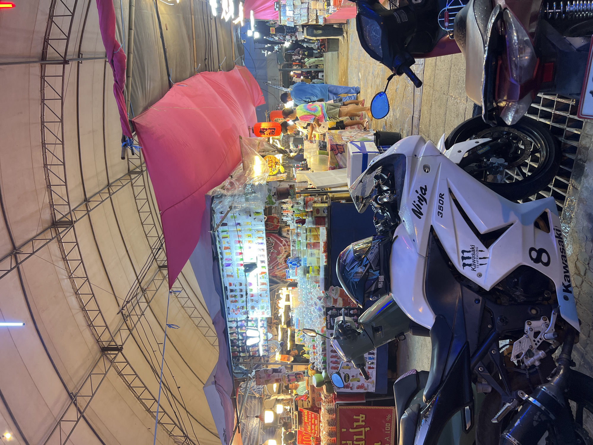 The image captures a bustling market scene at night in an unidentified location. The focus is on a white Kawasaki Ninja 300R motorcycle parked in the foreground, with another motorcycle visible behind it. A red canopy is draped overhead, and beneath it, vendors are selling various items, including beverages in numerous flavors displayed on shelves. People are browsing and purchasing goods. The market is lit by overhead fluorescent lights and the colorful displays of the stalls. The structure above the market consists of a metallic grid supporting a canvas roof.